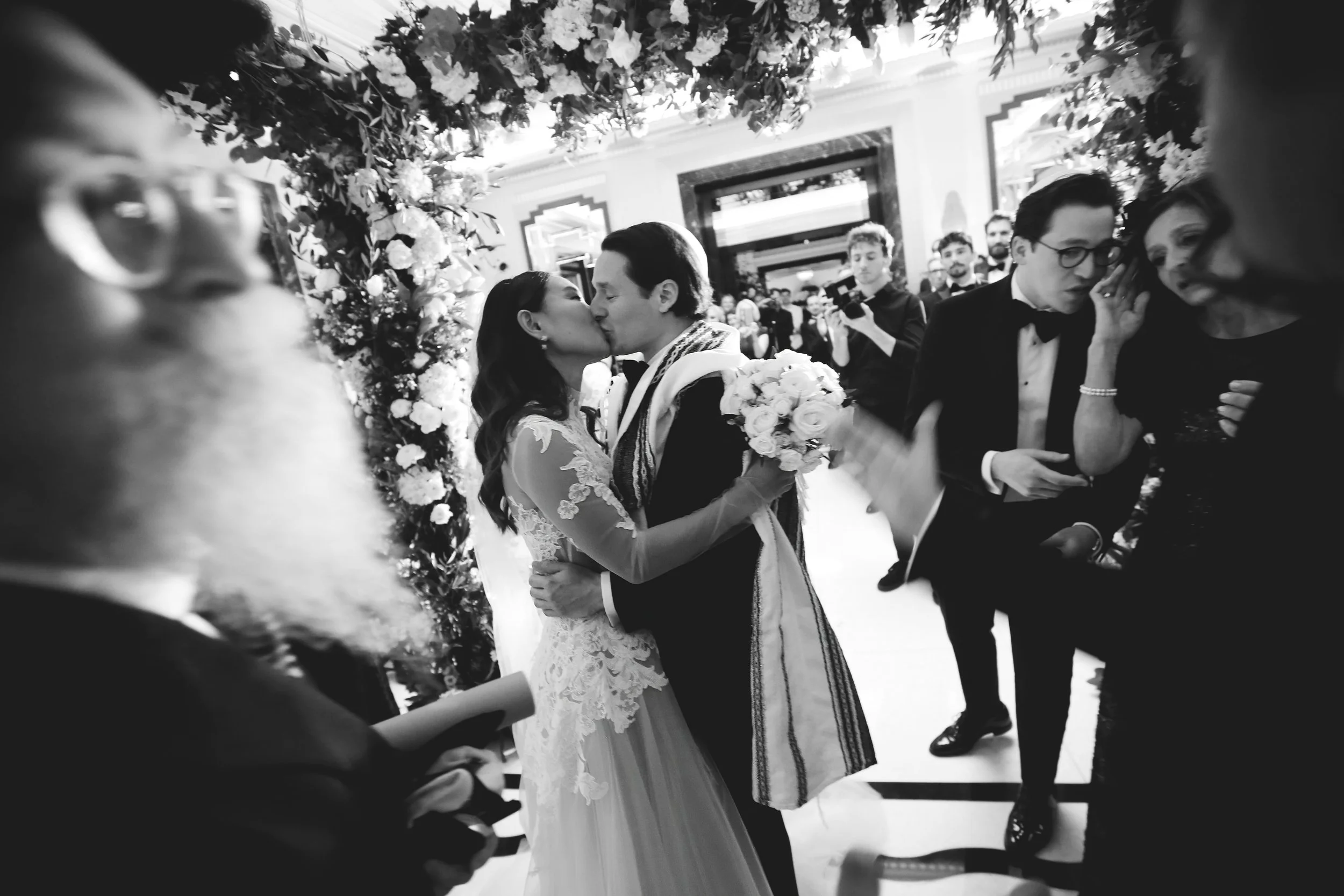 Bride and groom kissing under floral arch at a wedding ceremony with guests and photographer in the background.