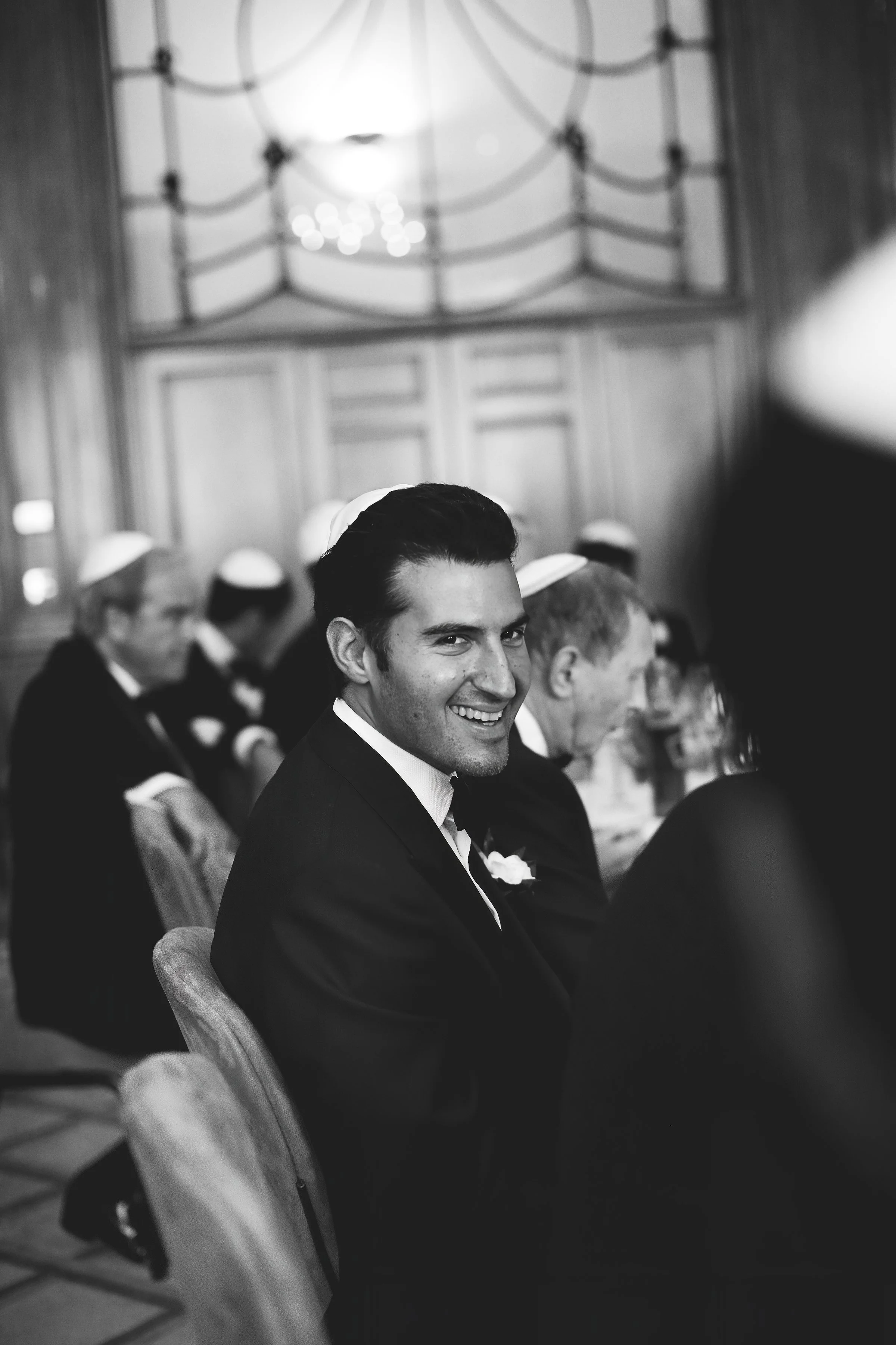 Black and white photo of a man in a suit smiling at an event, surrounded by people wearing kippahs.