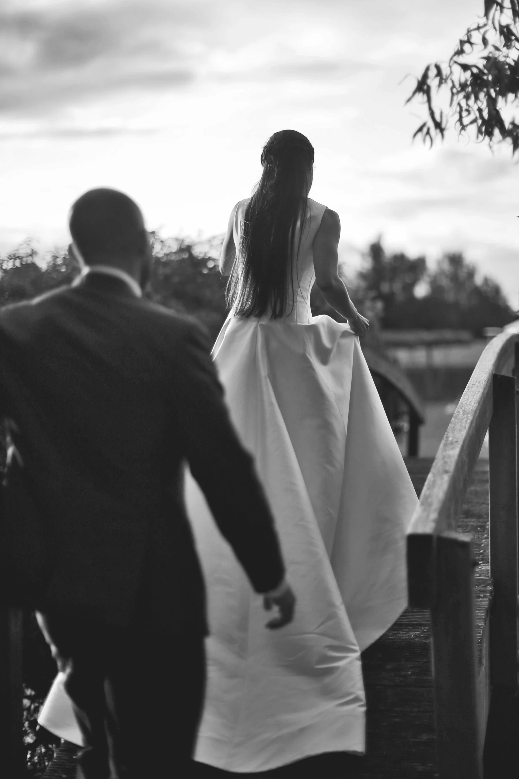 Bride walking on a bridge in a wedding dress, followed by a groom in a suit, black and white photograph.