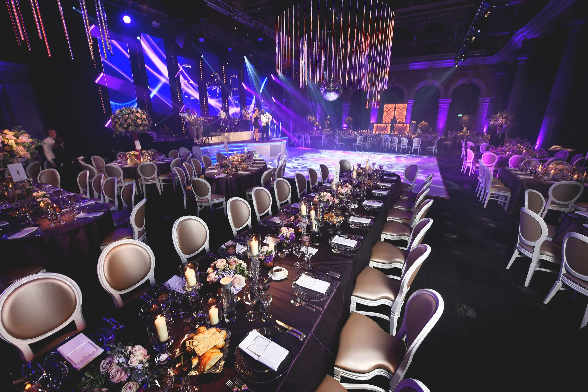 Elegant banquet hall setup with long dining tables, decorated with candles and floral centerpieces, under dramatic lighting. A stage with performers is visible in the background. The ambiance is luxurious and formal, suitable for a wedding or gala ev