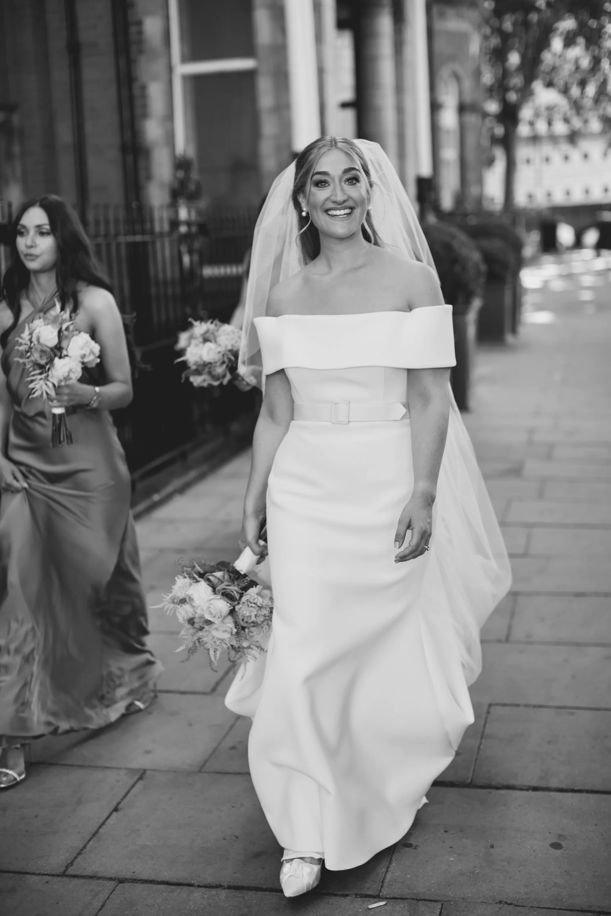 Bride in off-the-shoulder wedding dress holding bouquet, walking outdoors with bridesmaid, both smiling.