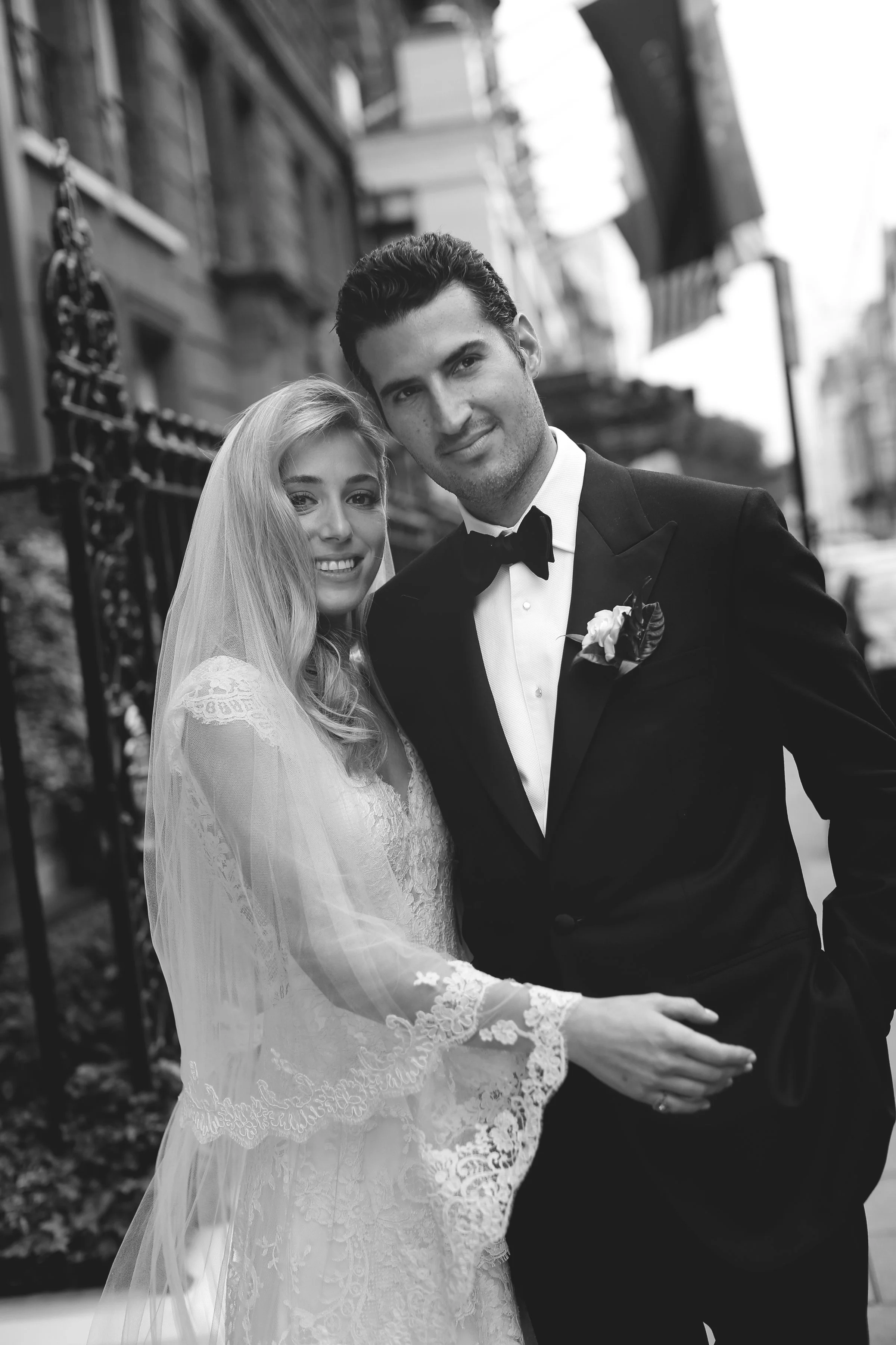 Black and white photo of a newlywed couple. The bride is wearing a lace wedding dress with a veil, and the groom is in a black tuxedo with a bow tie. They are standing outdoors with a building in the background.