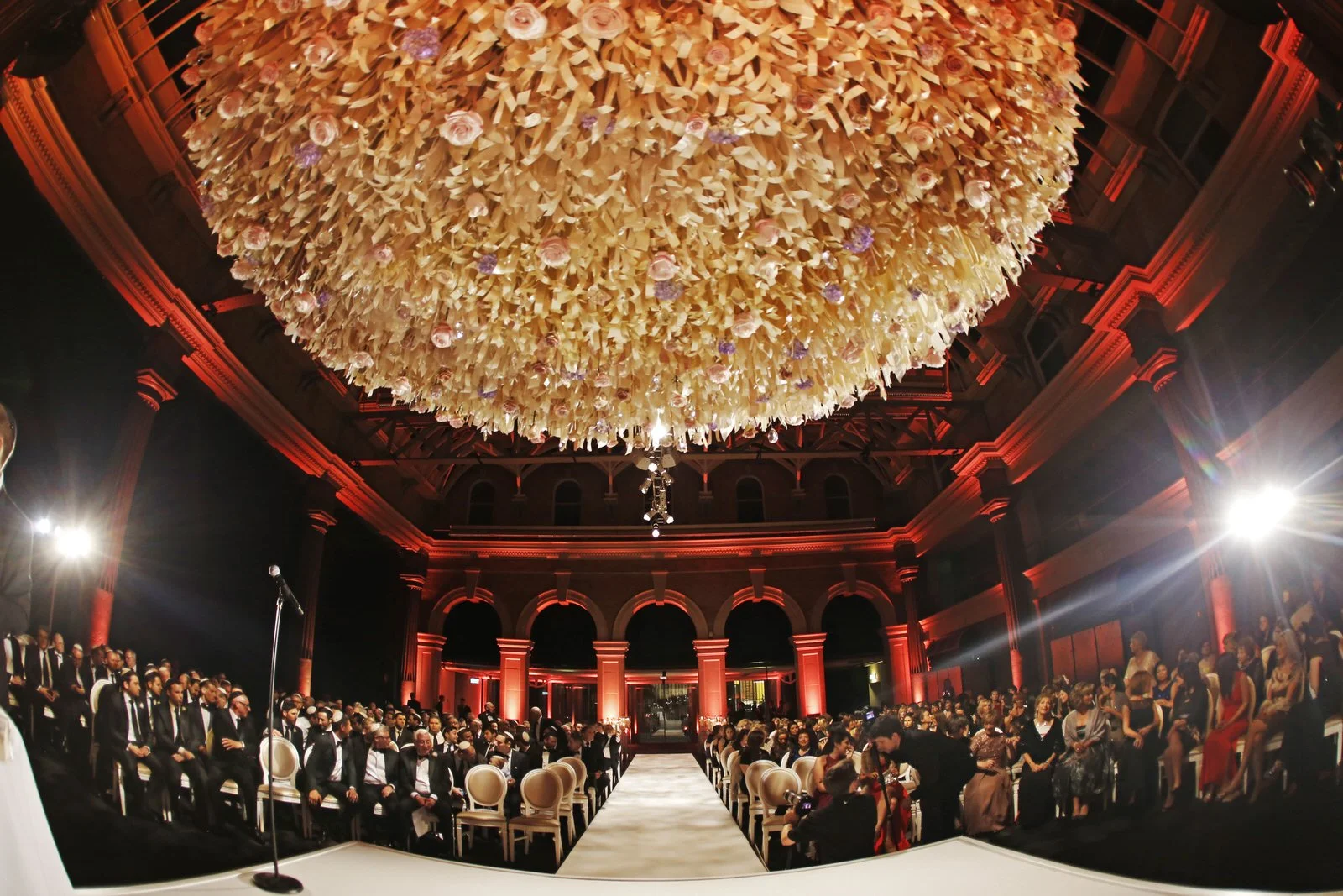 Elegant indoor event with a large, ornate floral chandelier hanging from the ceiling, surrounded by illuminated columns and a gathered crowd of formally dressed people, some seated and others standing, with a runway in the center.