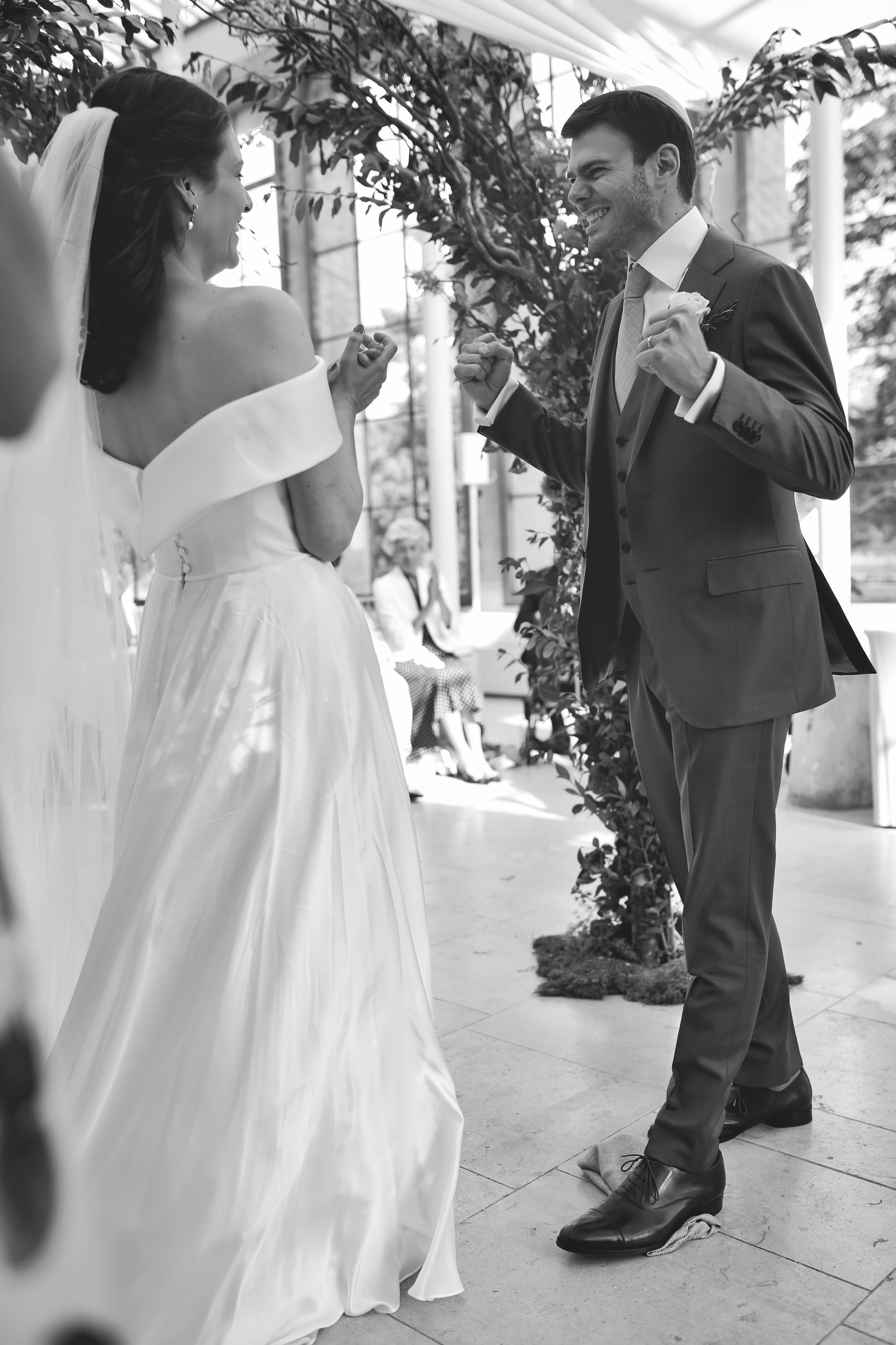 Bride and groom celebrating at wedding ceremony indoors, both smiling and facing each other, with a guest clapping in the background.