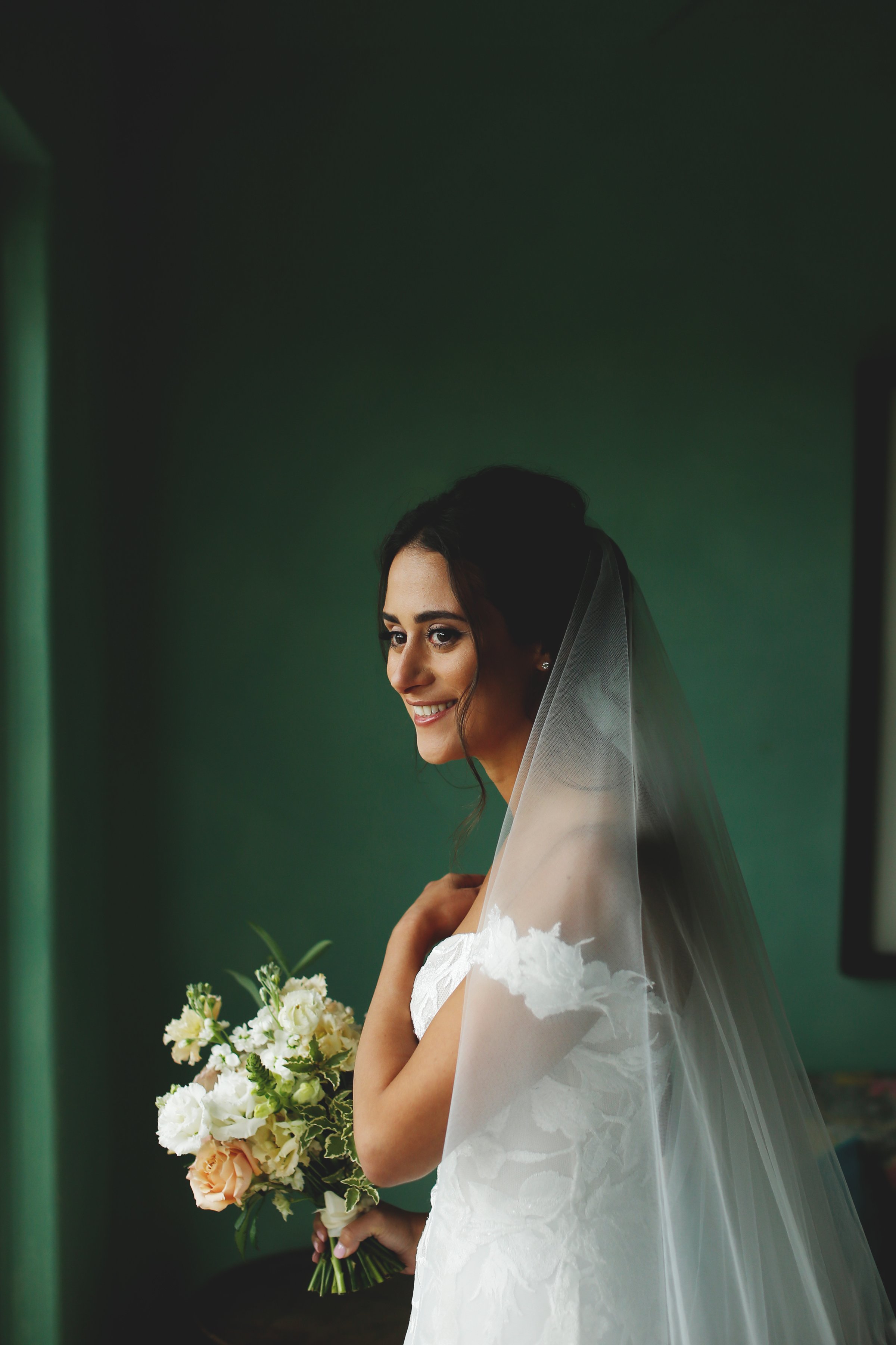 Bride in white dress holding a bouquet, smiling, standing near a window with a dark green background.