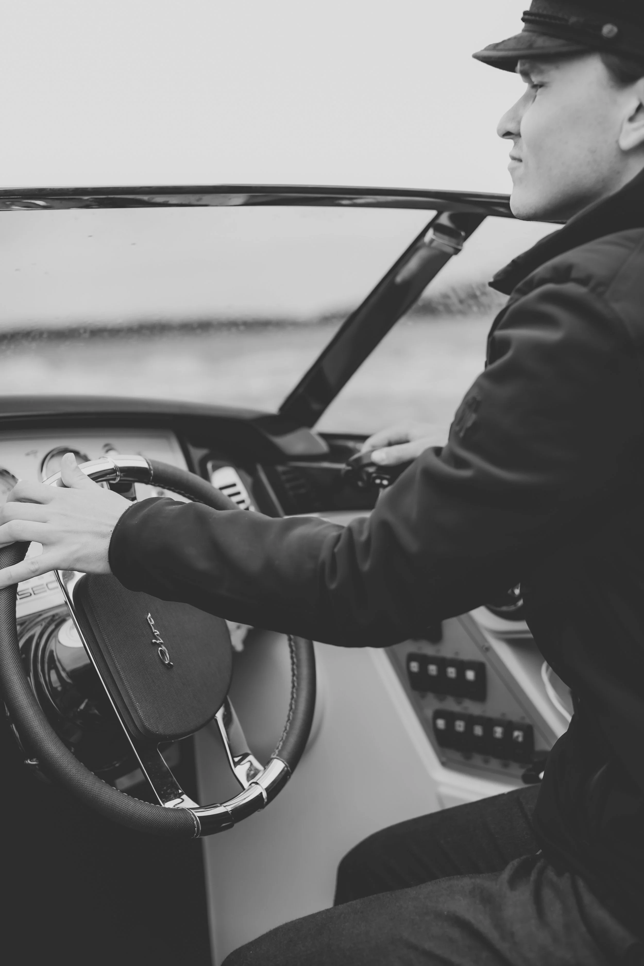 A person wearing a hat sitting in the driver's seat of a vintage car, holding the steering wheel, with a view of the dashboard and control panel visible.