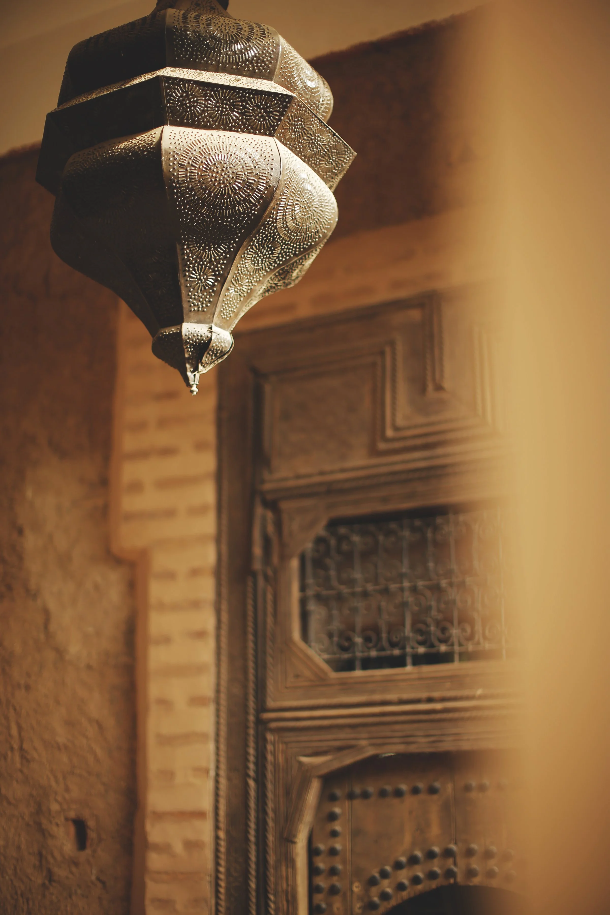 Close-up of an ornate metal hanging lamp with intricate circular and floral engravings, suspended above a traditional carved wooden door in a warmly lit room.