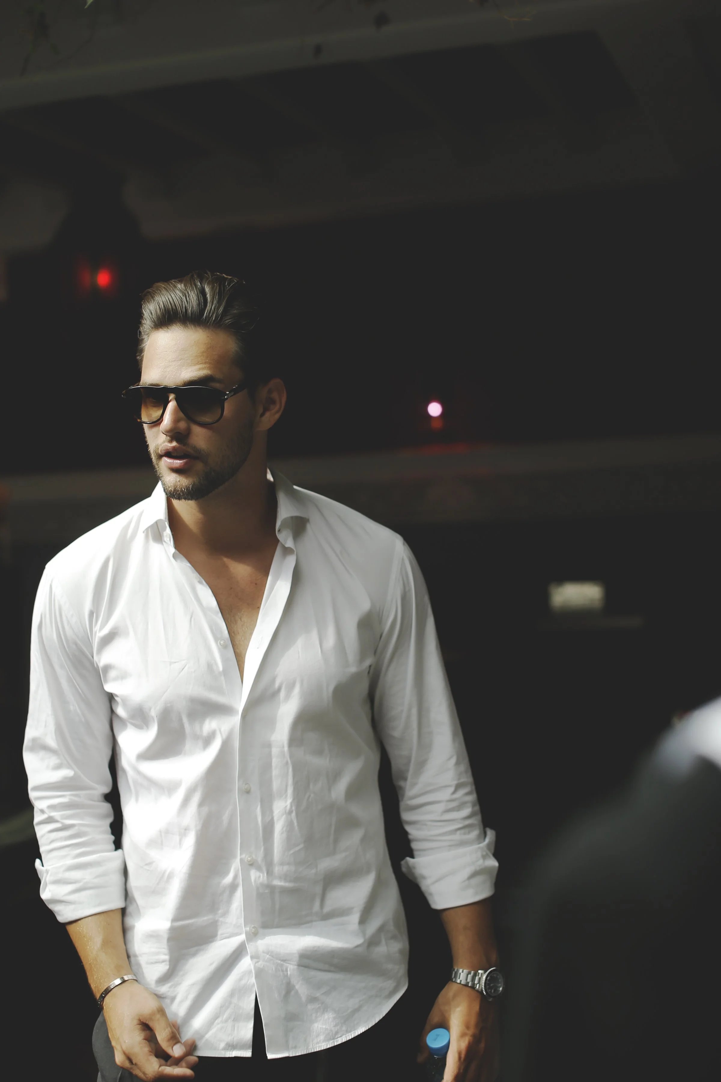 A man wearing dark sunglasses and a white button-up shirt with rolled-up sleeves, standing indoors against a dark background with faint colored lights, holding a water bottle in his right hand and wearing a watch and bracelet.