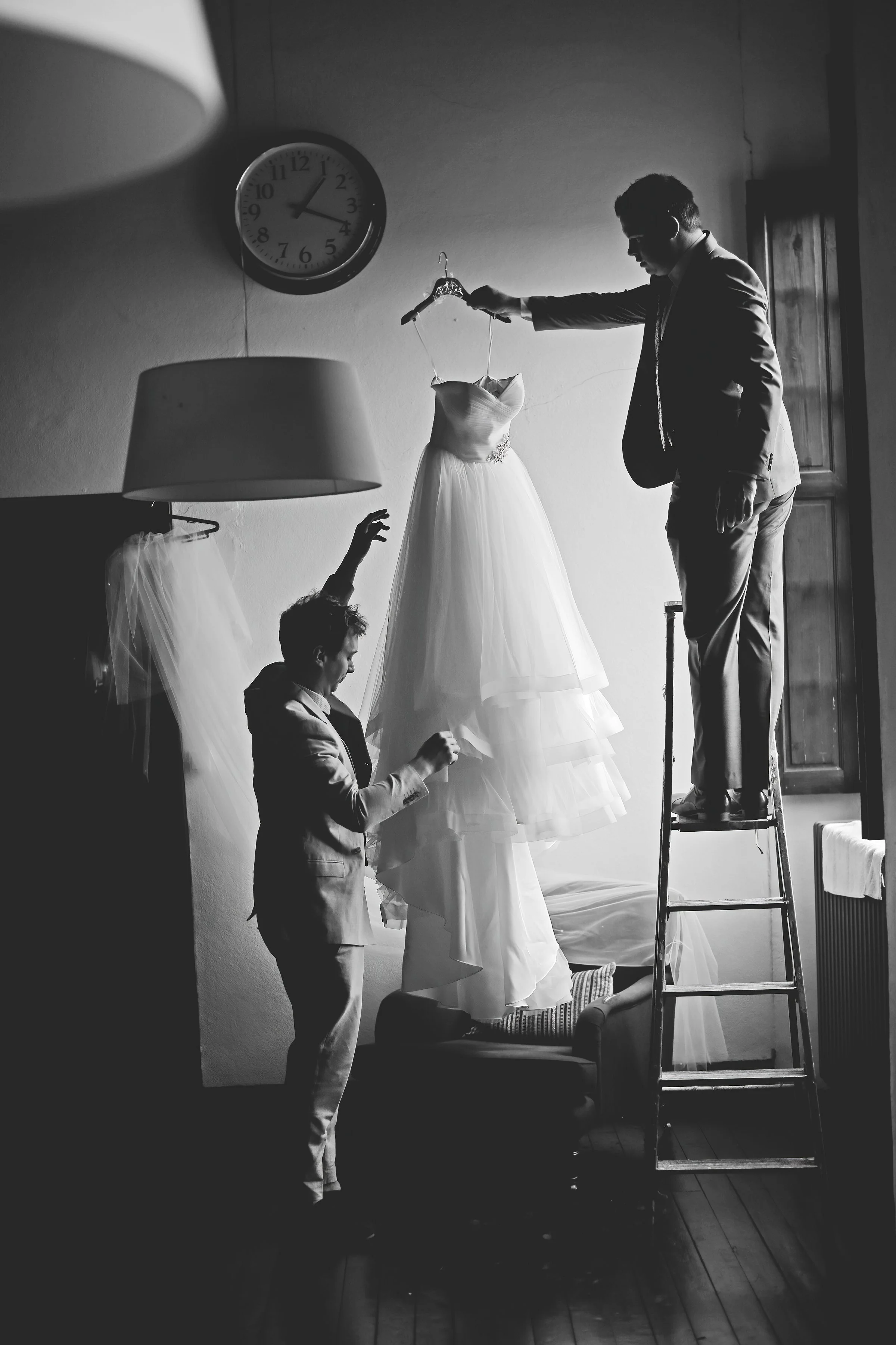 Black and white photo of two men arranging a wedding dress on a hanger, one standing on a ladder, with a vintage clock on the wall.