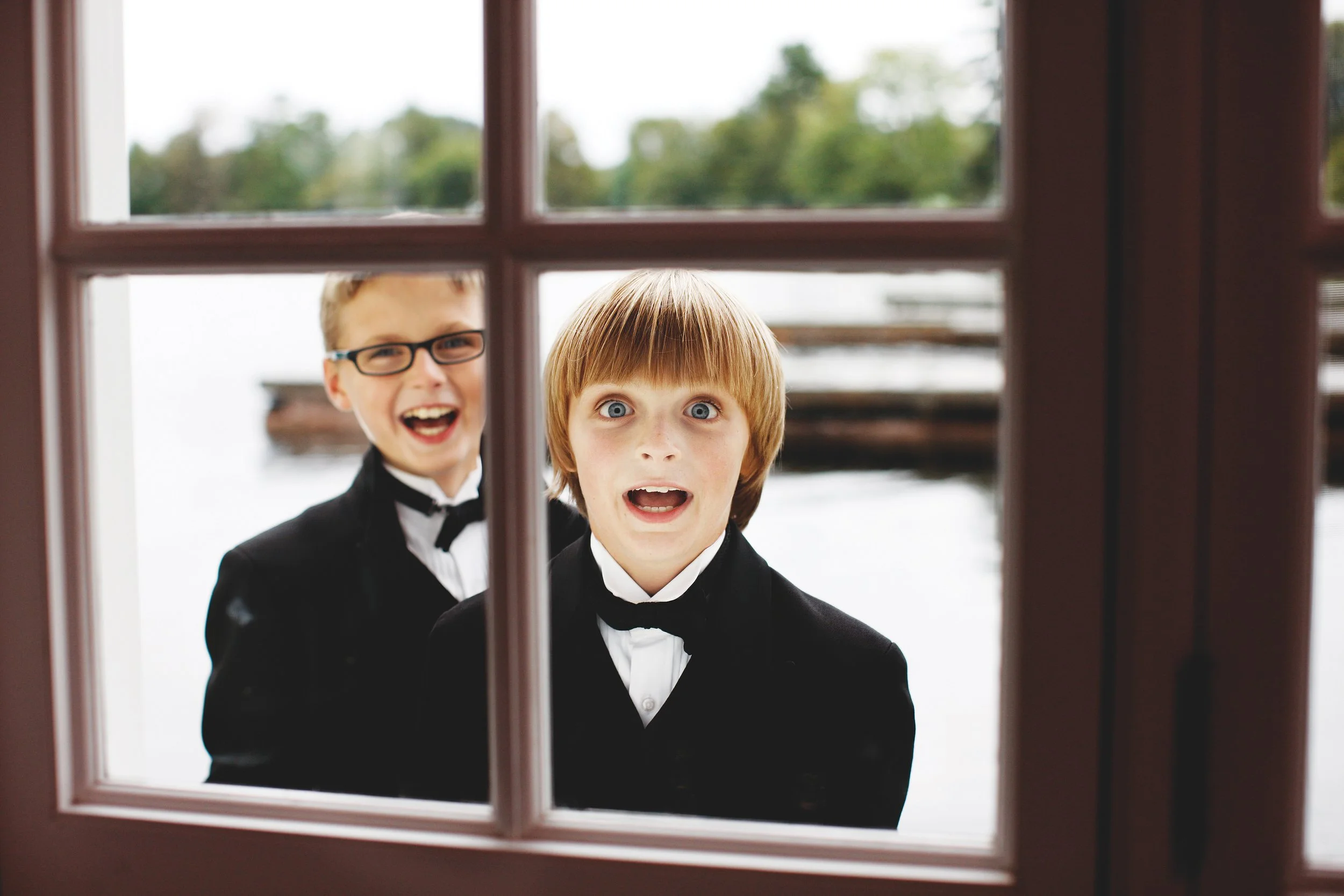 Two boys in suits looking through a window, one smiling and the other with a surprised expression.