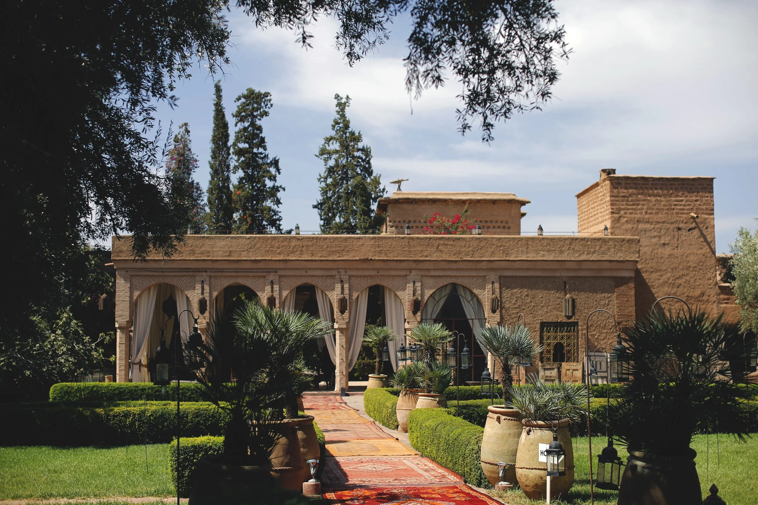 A building with arched windows and curtains, surrounded by lush green plants, trees, and potted plants, leading to a patio area with lanterns and a red carpet. Tall trees and a blue sky with some clouds are in the background.