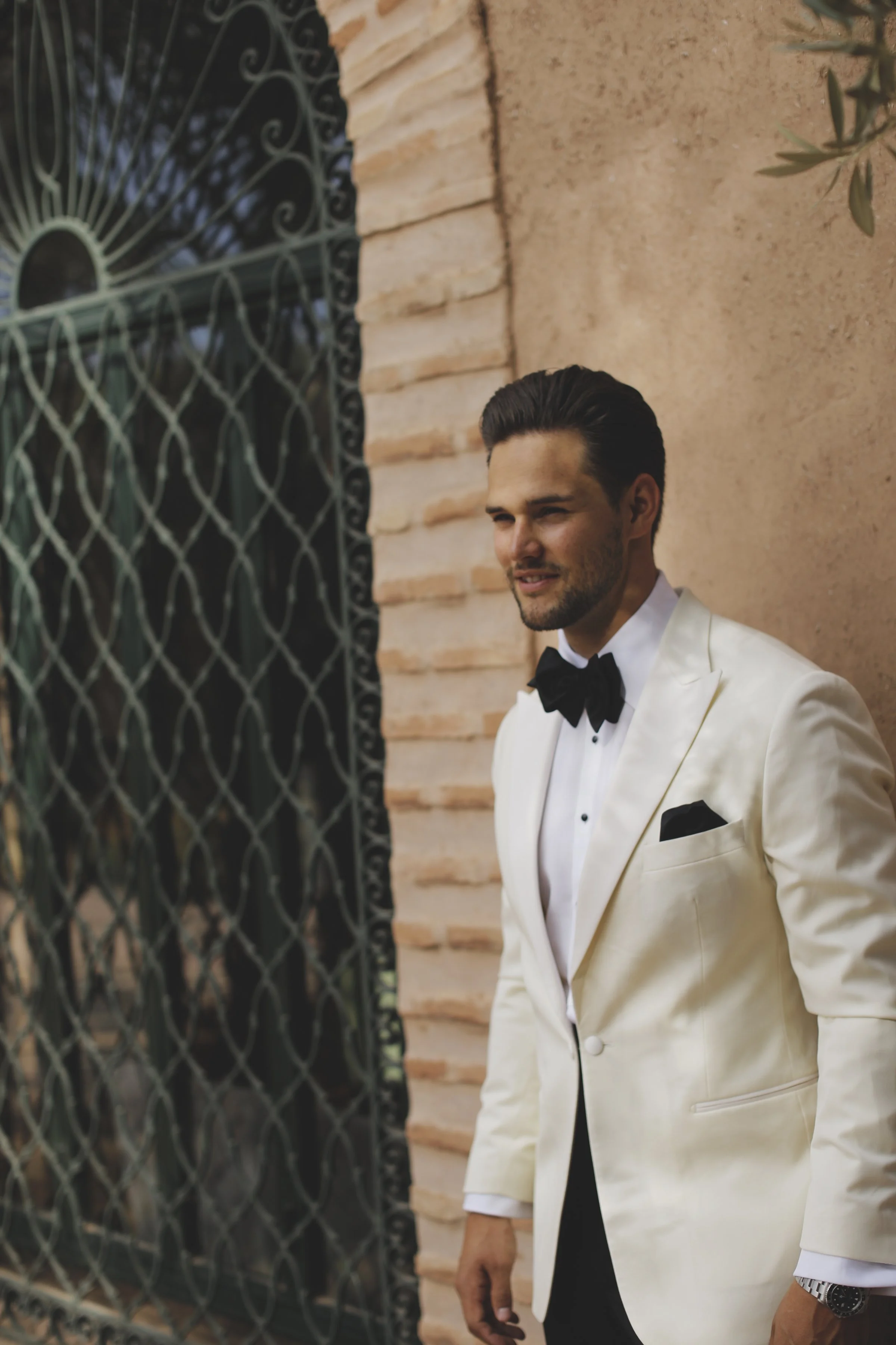 A man dressed in a white tuxedo with a black bow tie, standing next to a brick wall and a decorative metal gate.