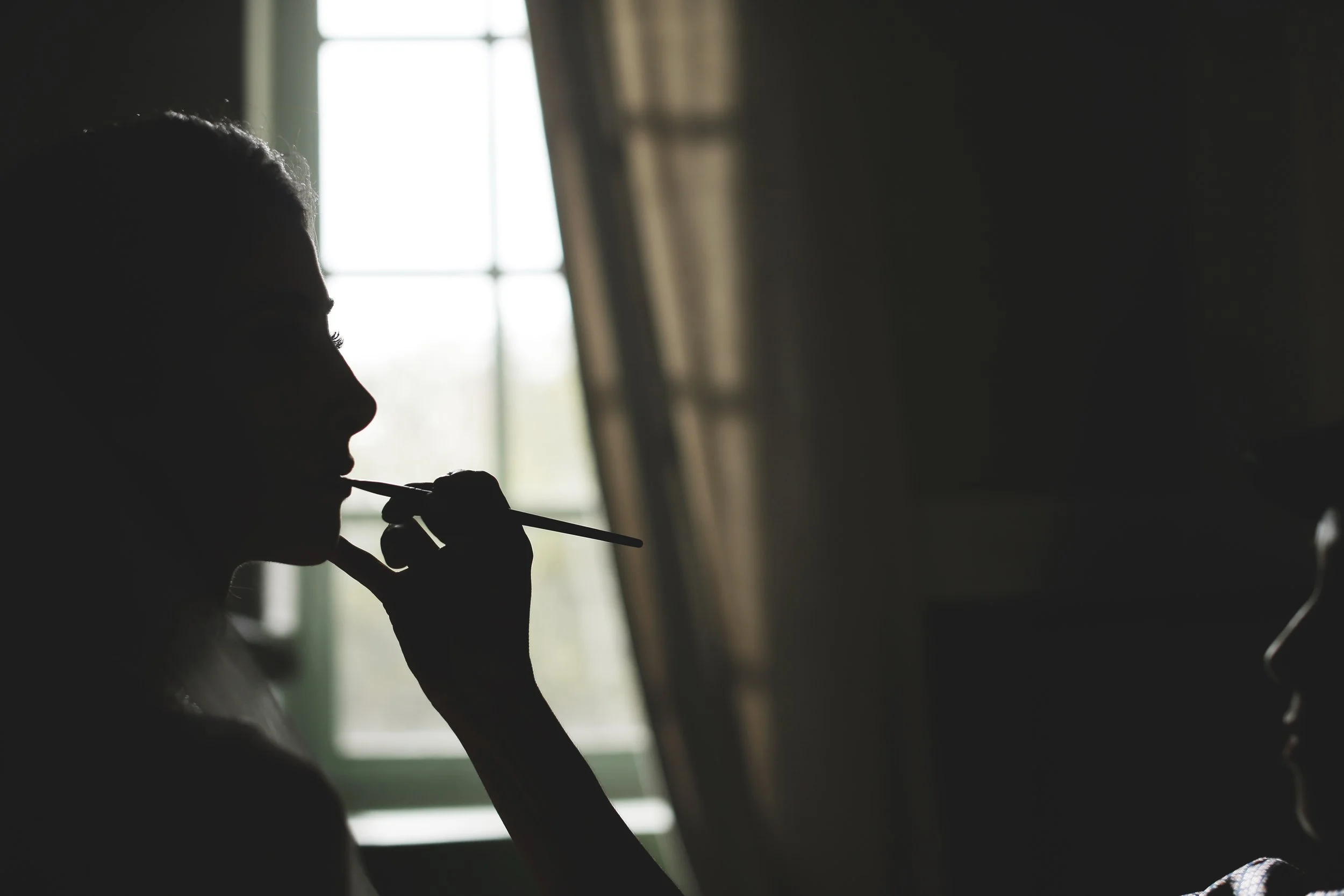 Silhouette of a person applying makeup in front of a window with curtains, backlit by natural light.