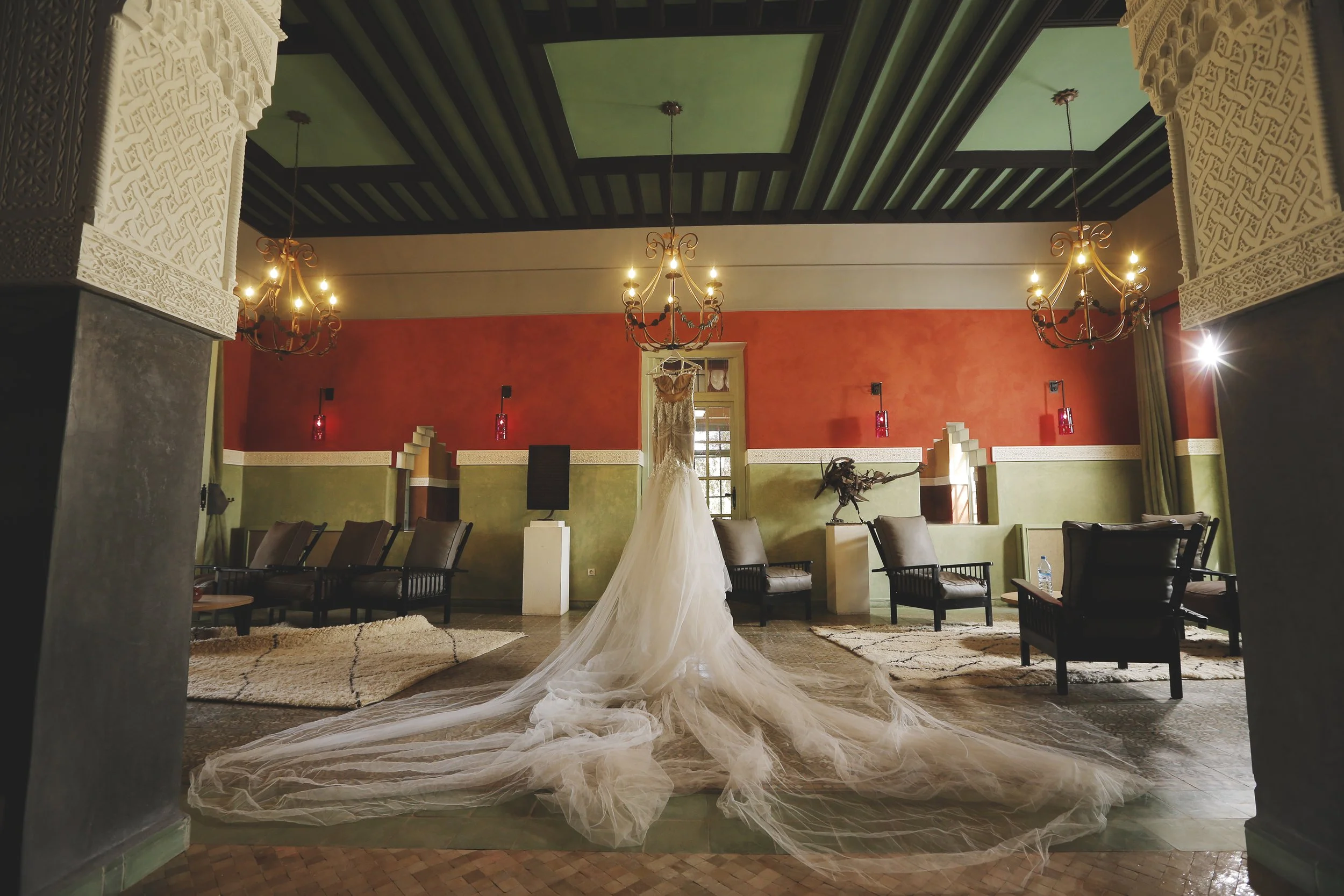 An elegant wedding dress on a mannequin in a decorated room with chandeliers, red walls, and green ceiling.