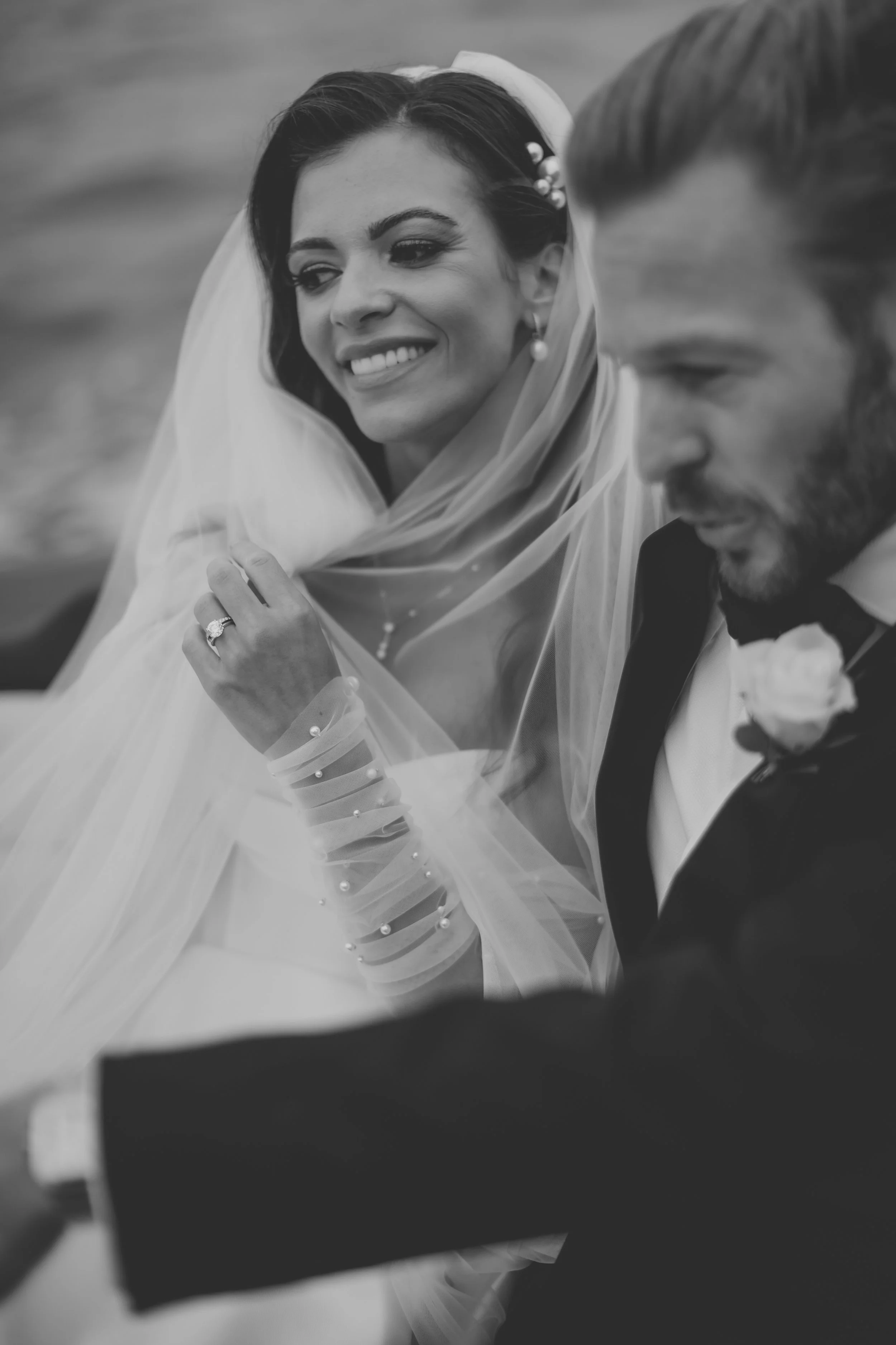 A black and white photograph of a bride and groom. The bride has dark hair with floral accessories and is smiling, while the groom has light hair and is dressed in a tuxedo. They appear to be at their wedding.