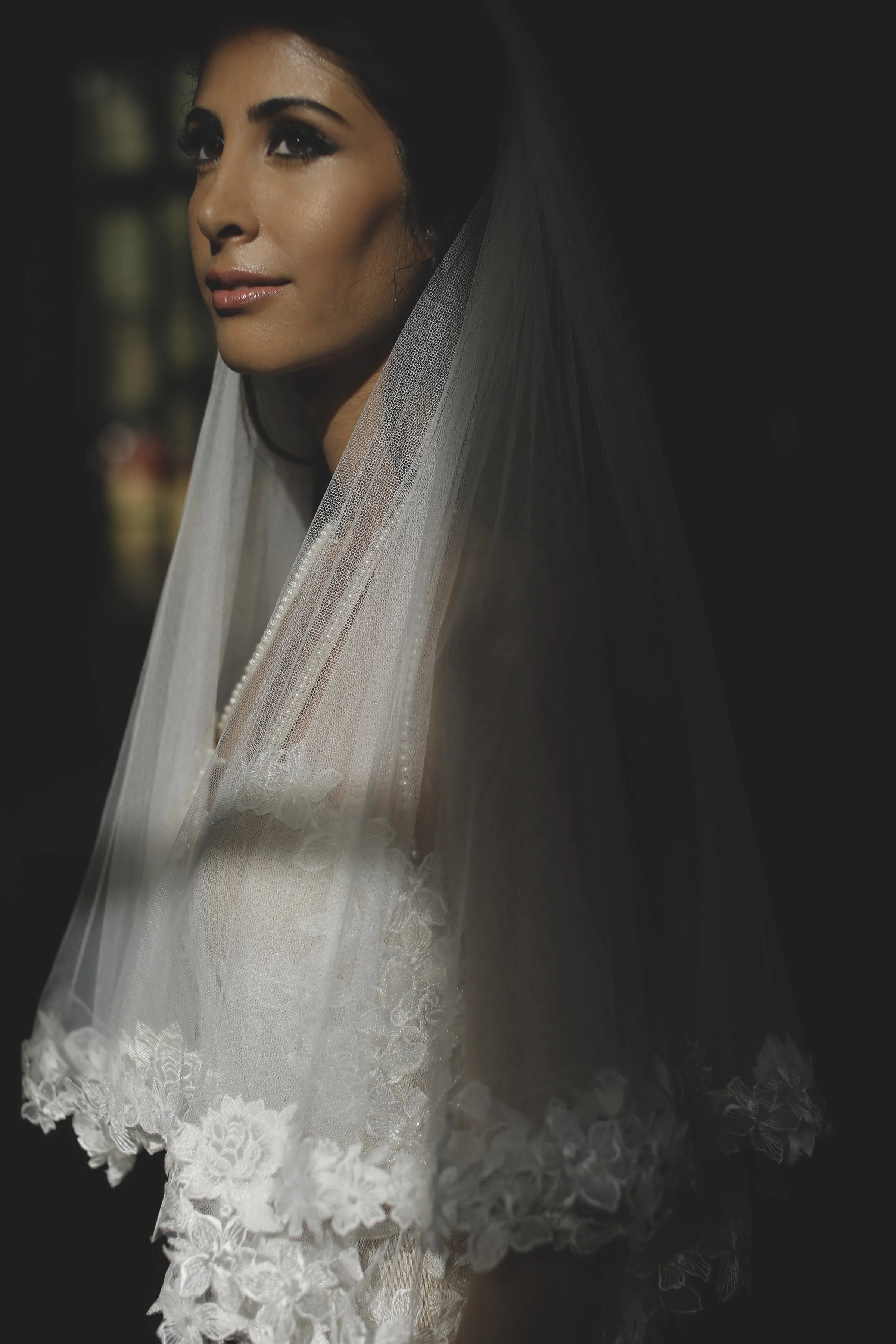 A woman wearing a wedding dress with lace details and a veil, looking slightly upwards, in a dimly lit setting.