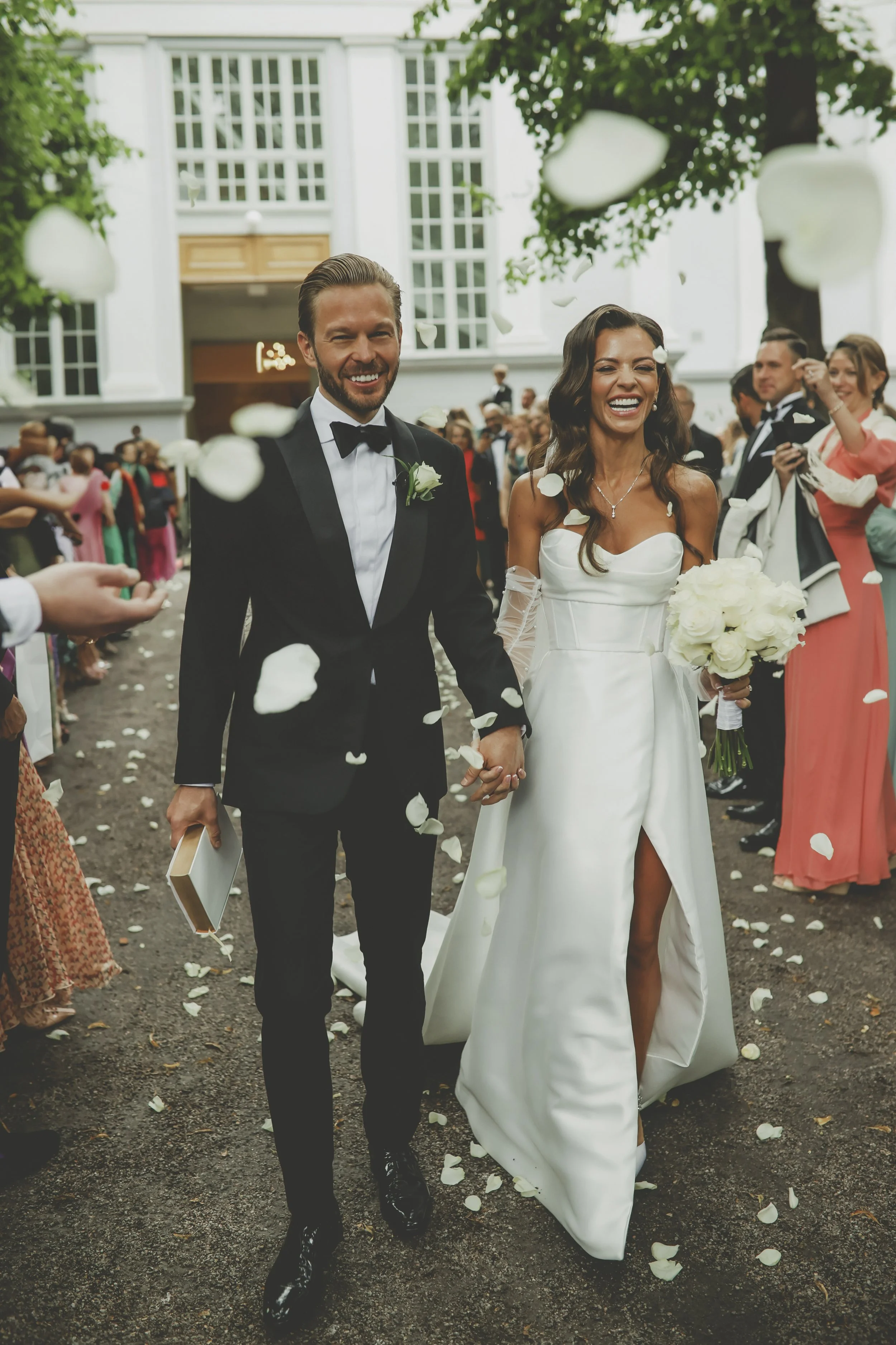 A joyful bride and groom walking hand in hand after their wedding ceremony surrounded by guests throwing flower petals outside a grand white building.