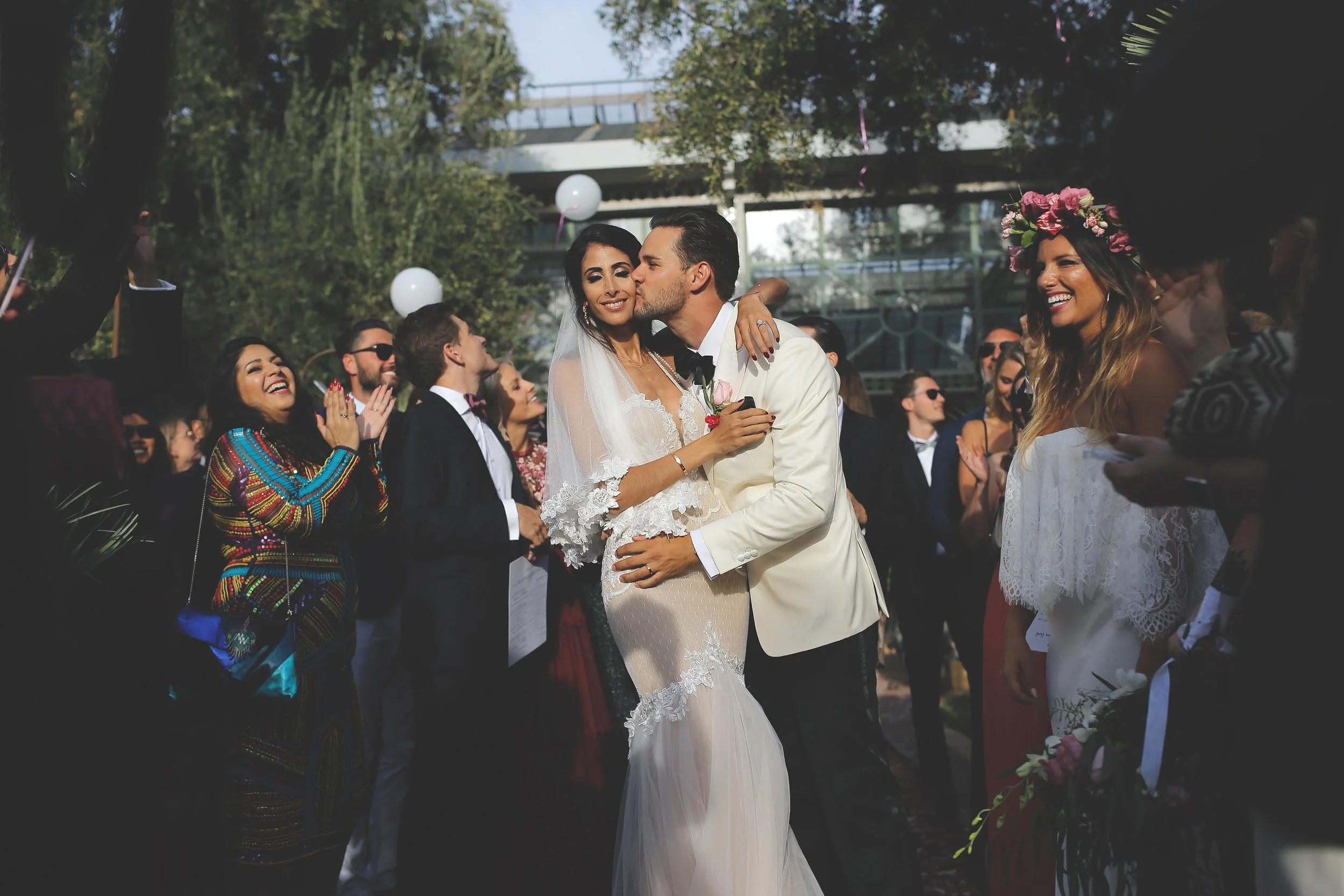 Bride and groom kissing at outdoor wedding with guests cheering and smiling.