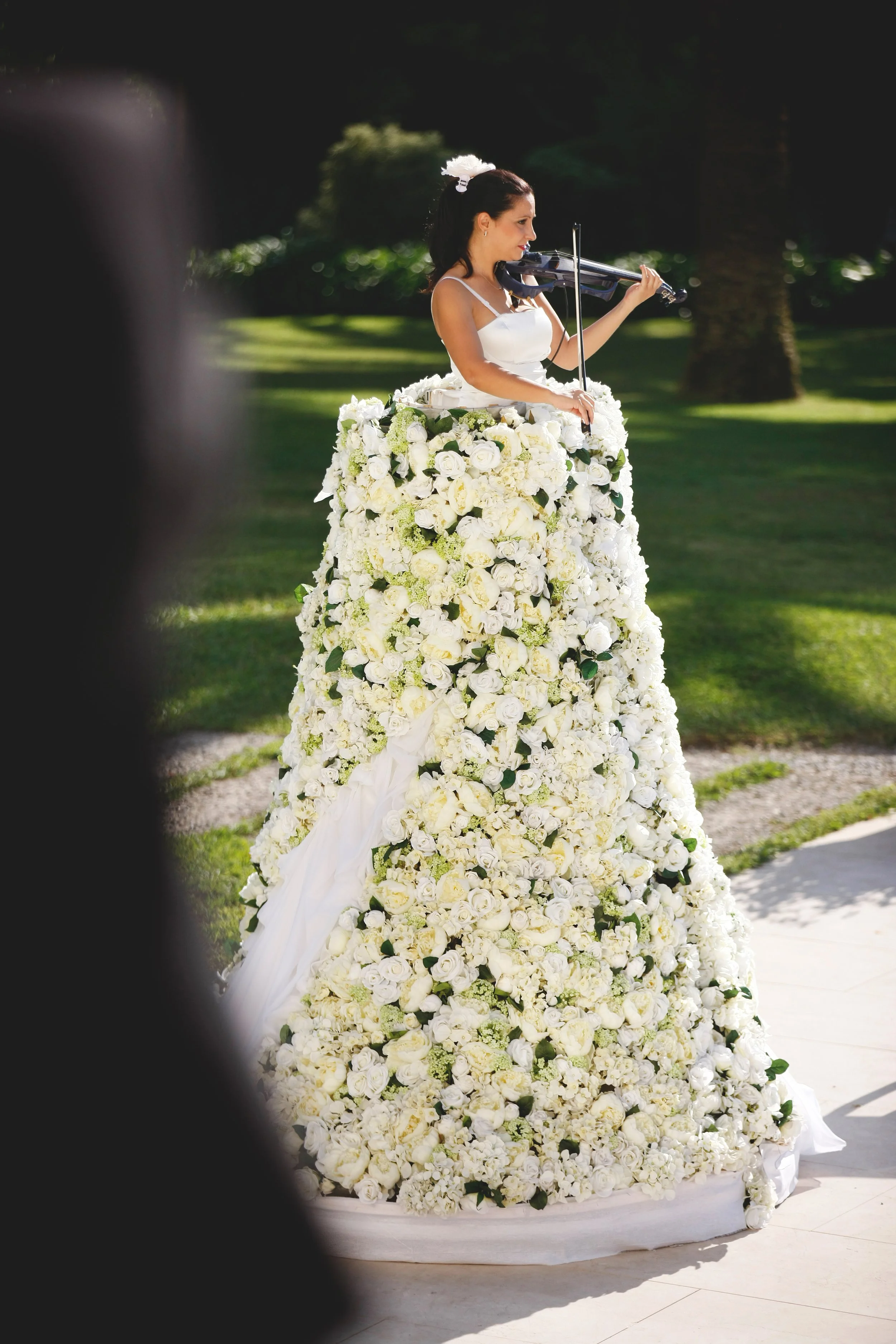 A woman standing inside a large, dress-shaped floral arrangement, playing a violin outdoors.
