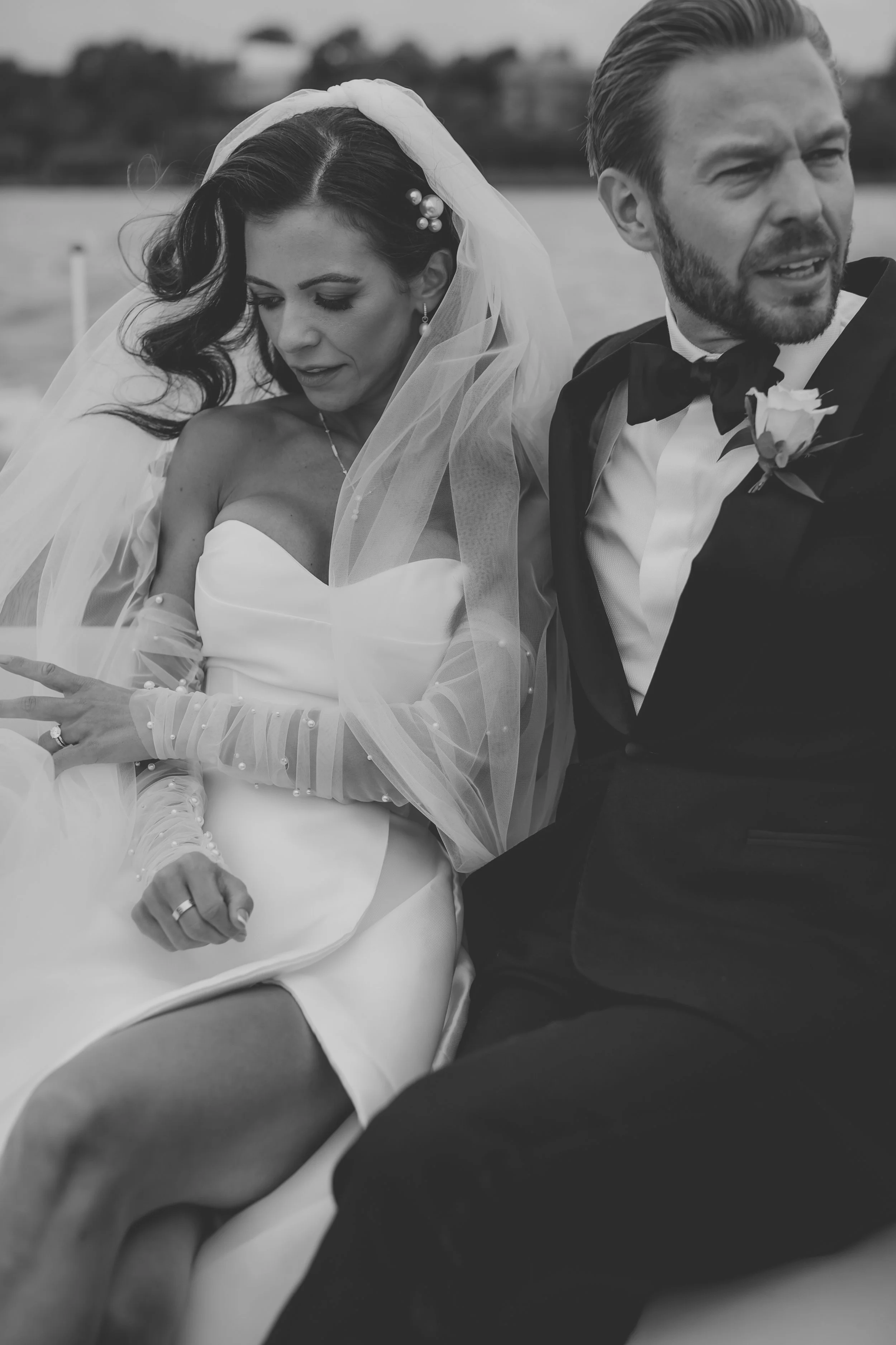 A black and white photo of a bride and groom sitting close together outdoors, with the bride looking down and the groom looking off to the side.