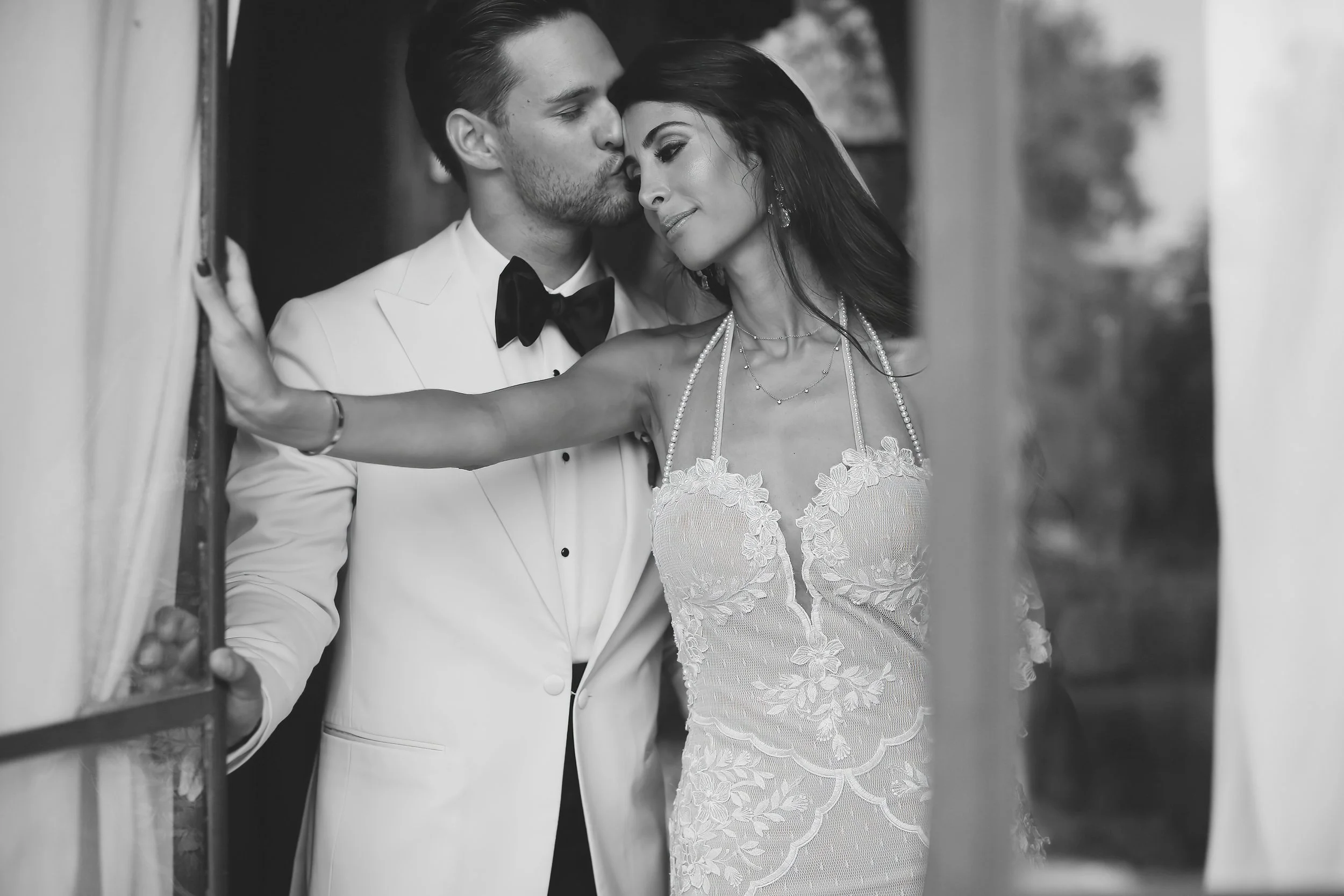 Black and white image of groom kissing bride's forehead, bride in lace gown, groom in white tuxedo.