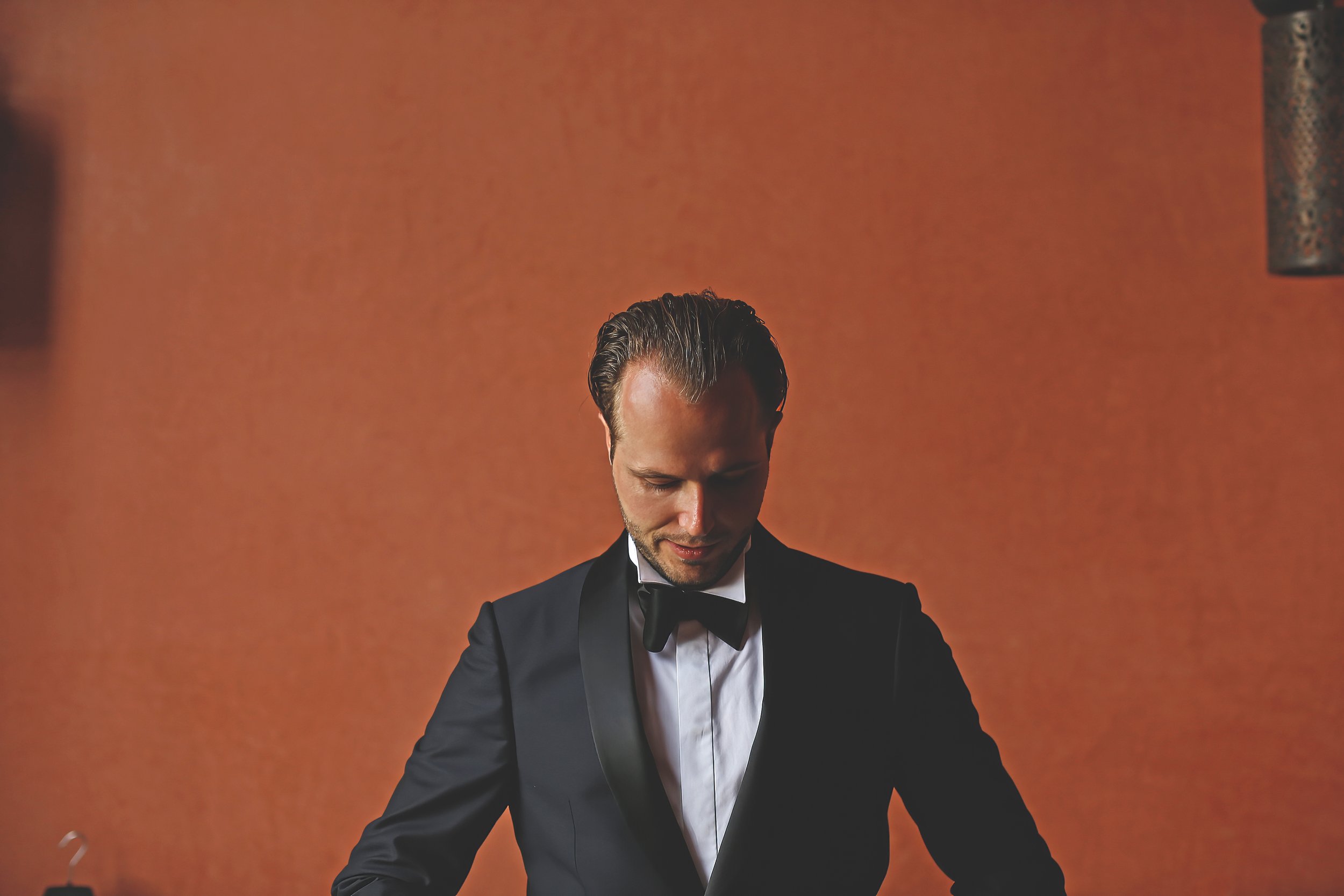 Man in formal suit with bow tie against orange background, looking down.