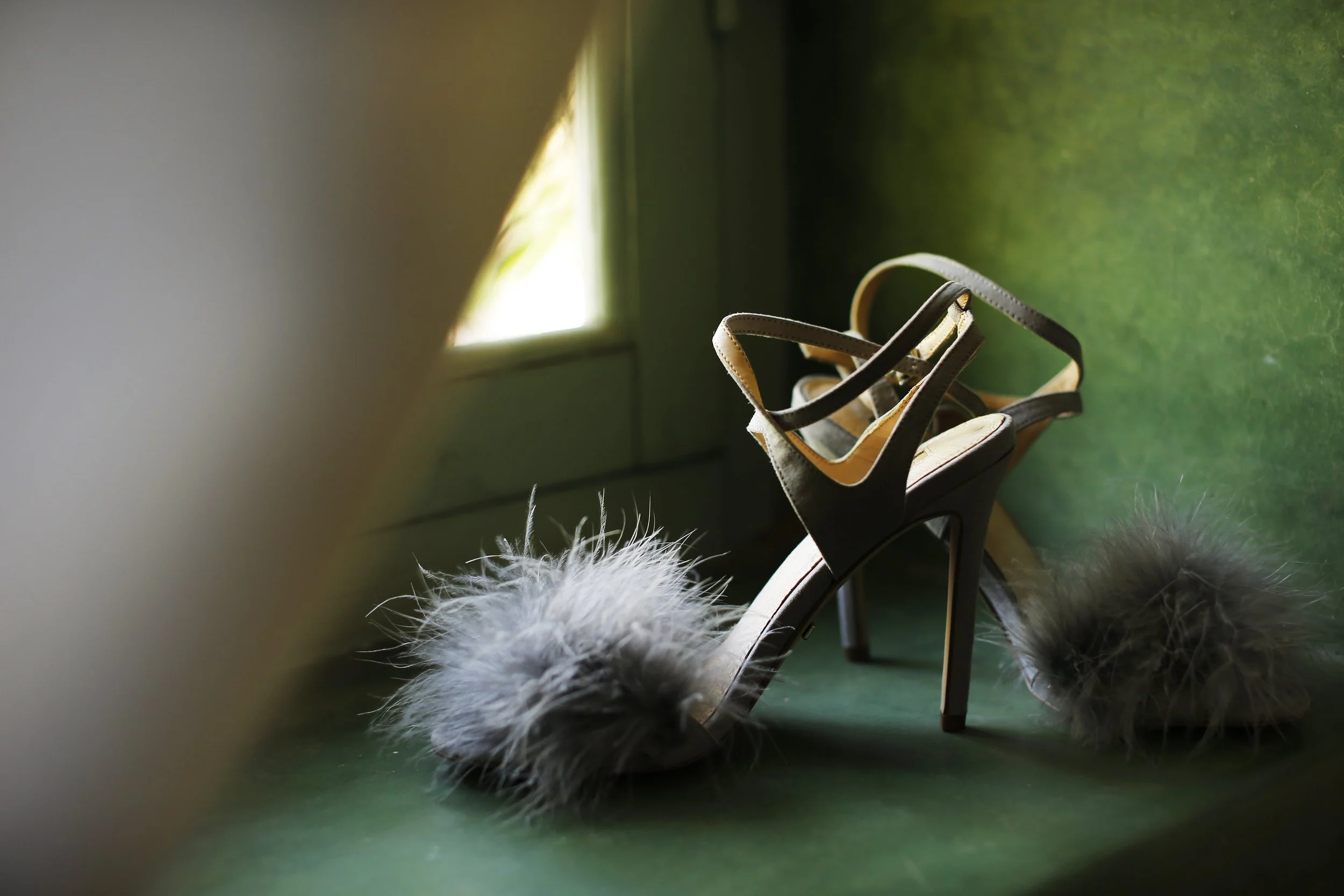 A pair of beige high-heeled sandals placed on a green surface near a window, with fluffy white and black fur at the edge of the frame.