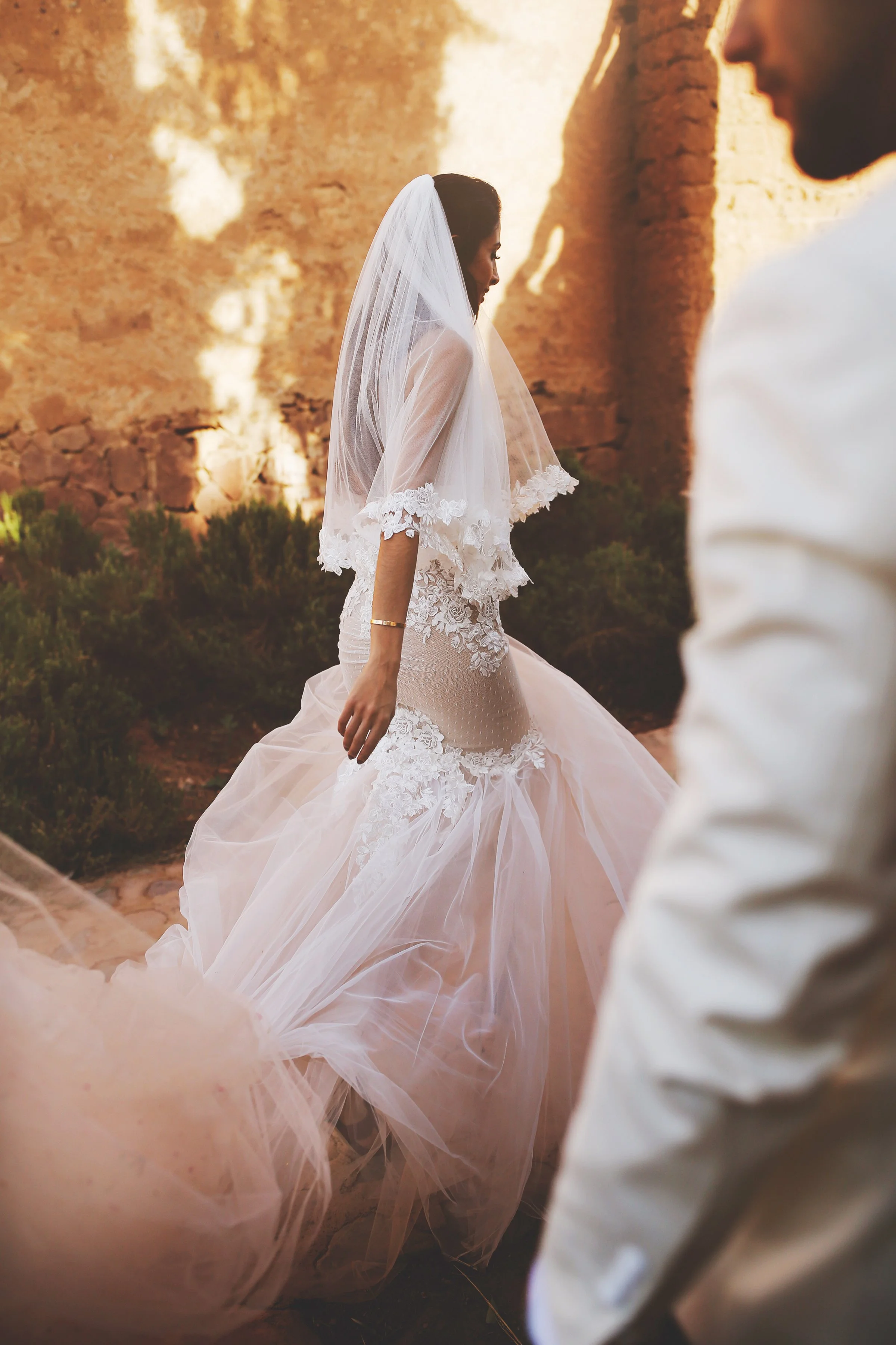 Bride in a wedding dress with a veil walking outdoors, accompanied by a groom in formal attire, with a rustic wall in the background.