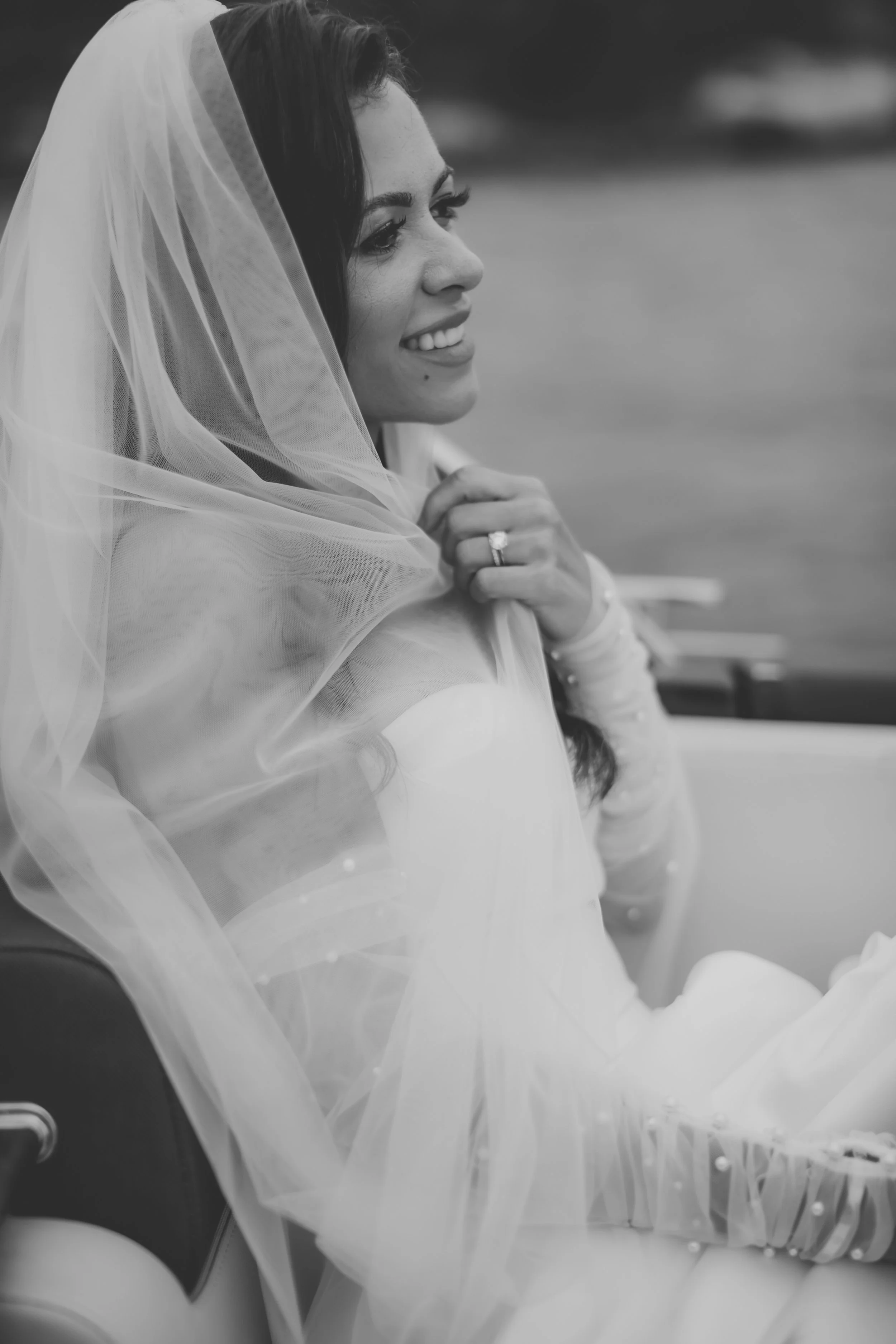 A woman in wedding attire smiling, wearing a veil and diamond ring, sitting outdoors.