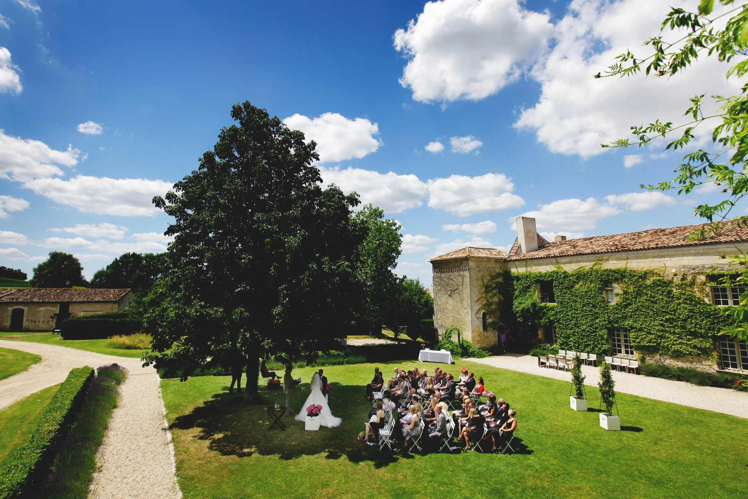 Outdoor wedding ceremony in a garden, guests seated, bride and groom under a tree, historic building with ivy in background, blue sky.
