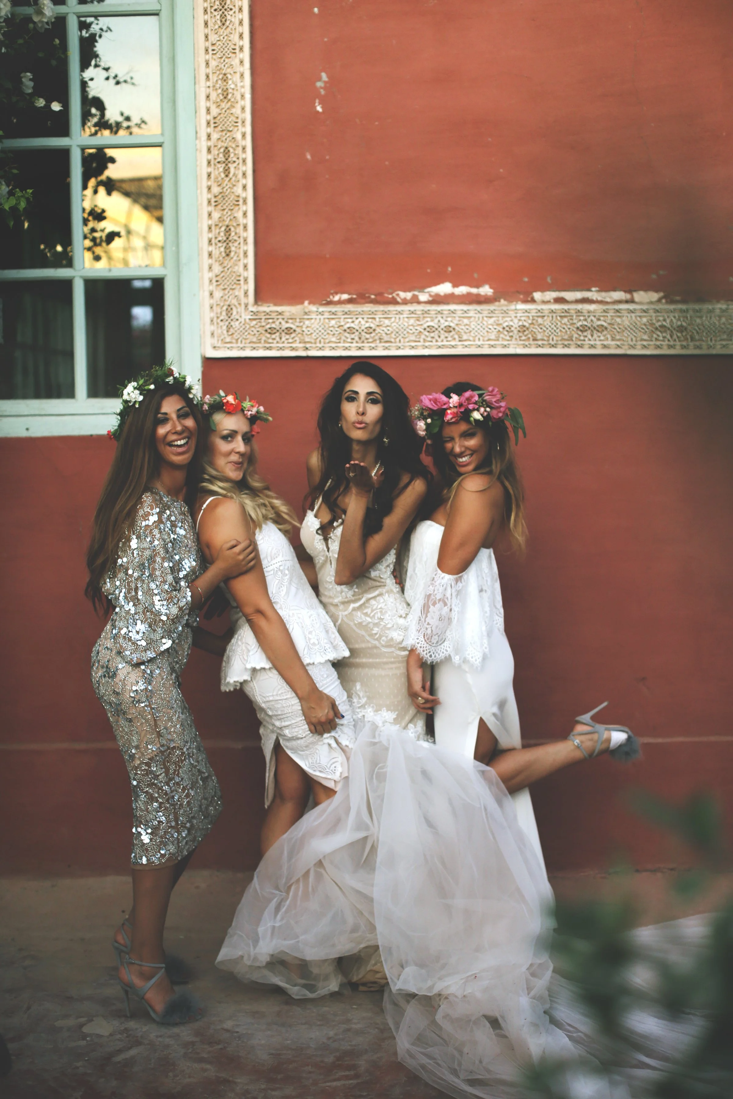Four women in wedding dresses and floral crowns posing playfully against a red wall with a window.