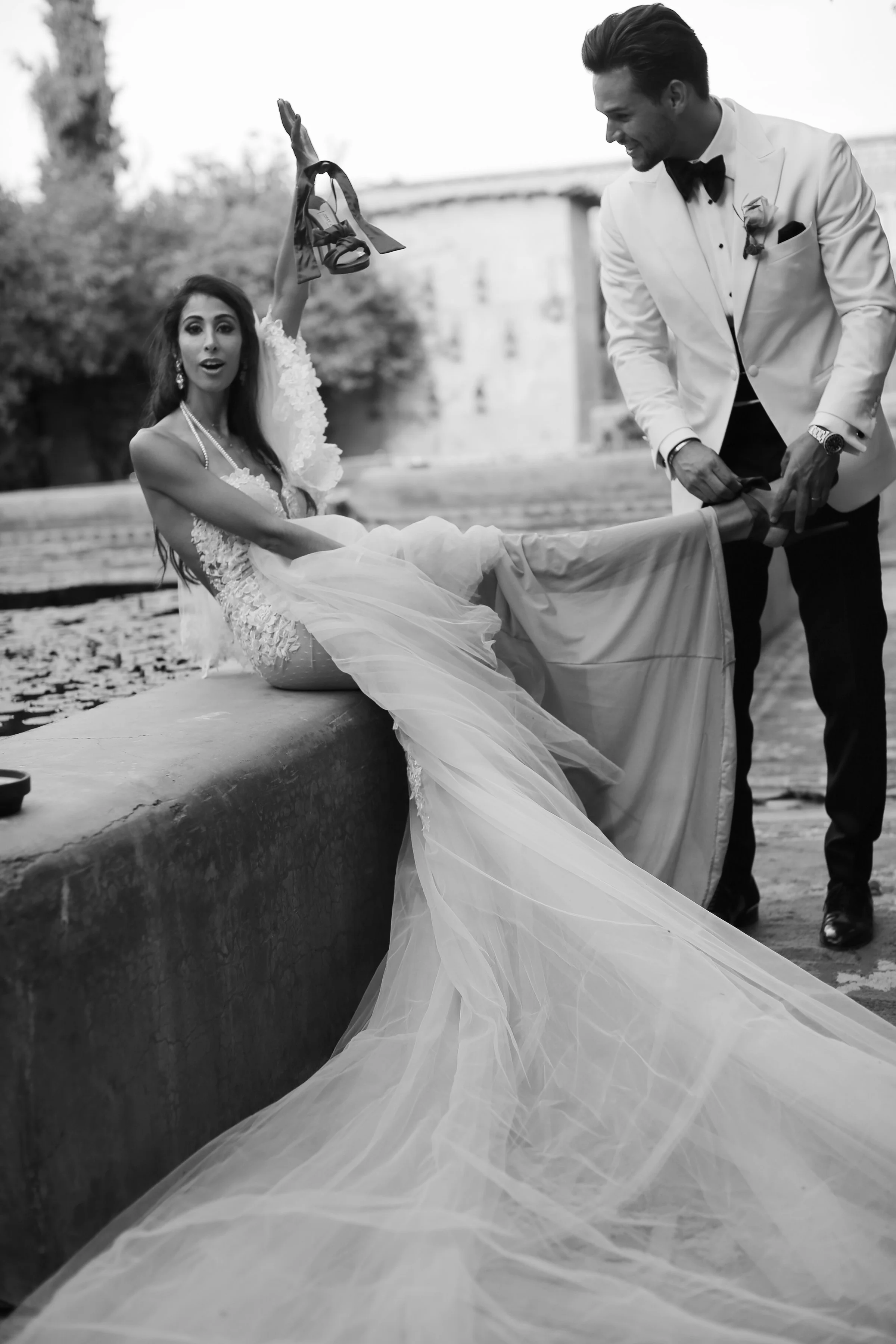 A bride and groom during their wedding photo shoot outdoors, with the bride sitting on a ledge and the groom bending over her, holding her shoe, while she holds up her shoe with a surprised expression. The bride wears a lace wedding dress, and the gr