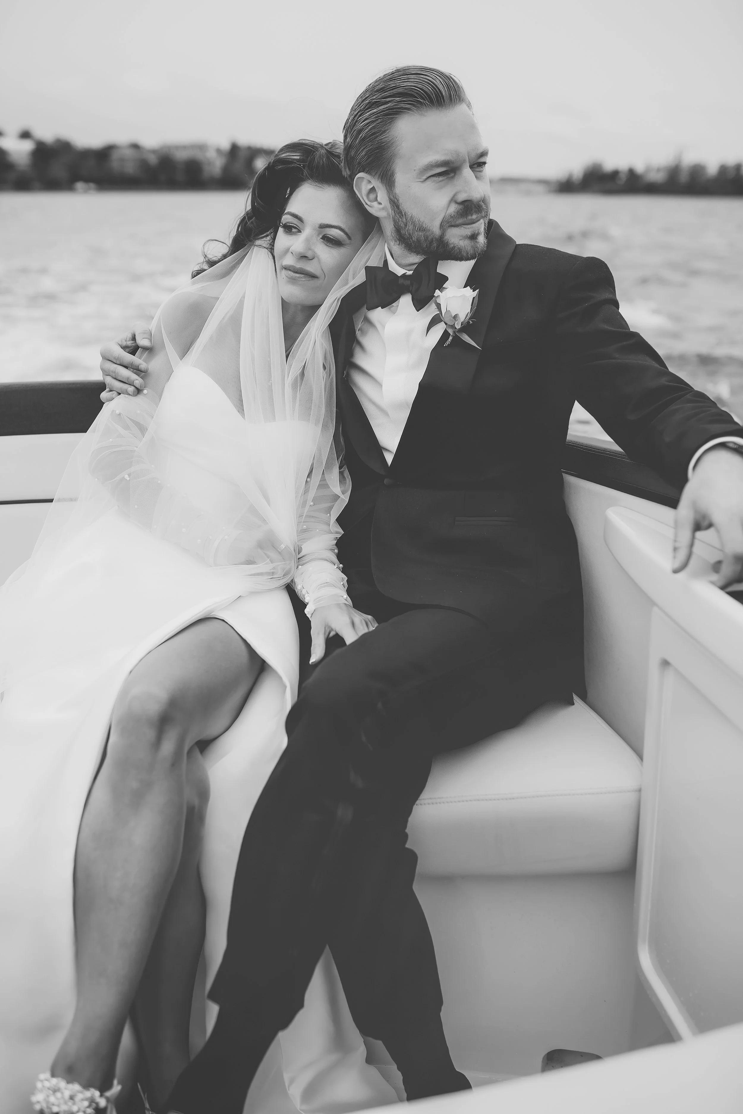Elegant couple on a boat, bride in a gown with veil, groom in a tuxedo, serene water background.