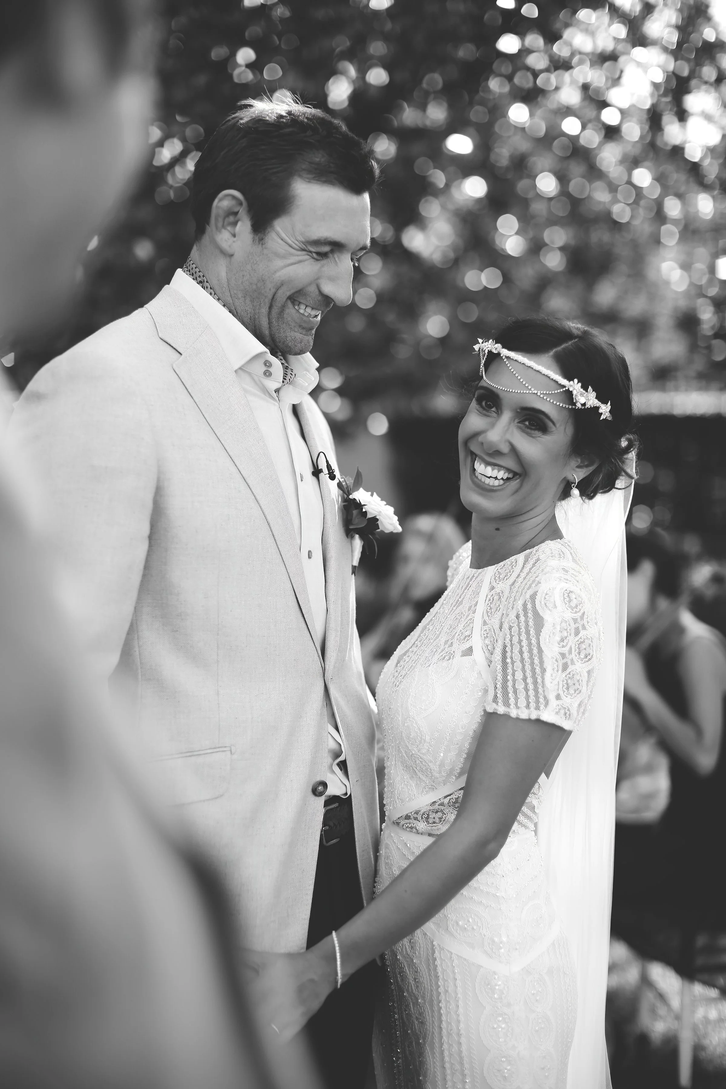 A black and white photo of a bride and groom smiling on their wedding day. The bride wears a lace dress and headpiece with a veil, while the groom is in a light-colored suit with a boutonniere.