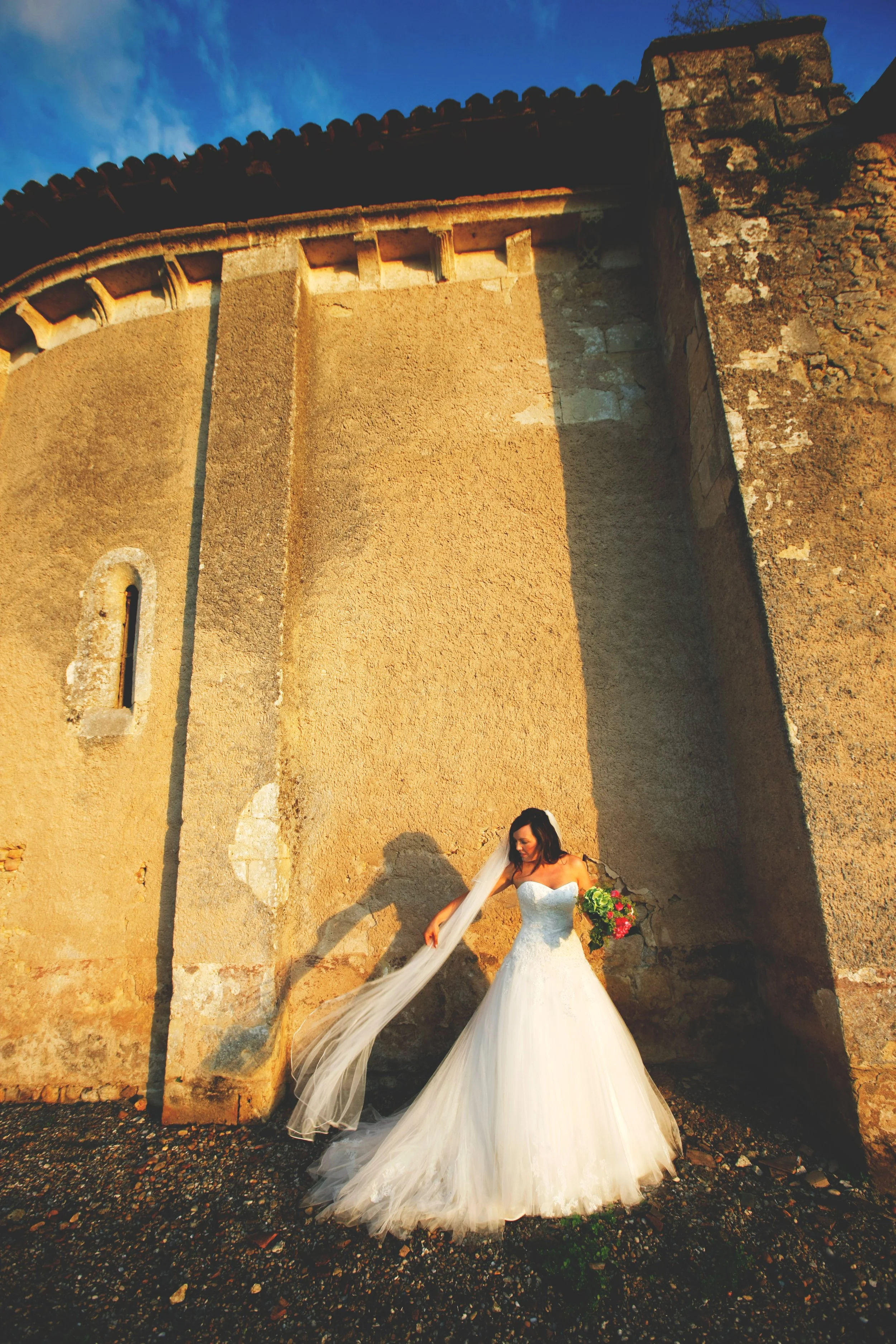 Bride in white wedding dress holding bouquet, standing against an old stone building wall with a small window; sunlight casts long shadows.