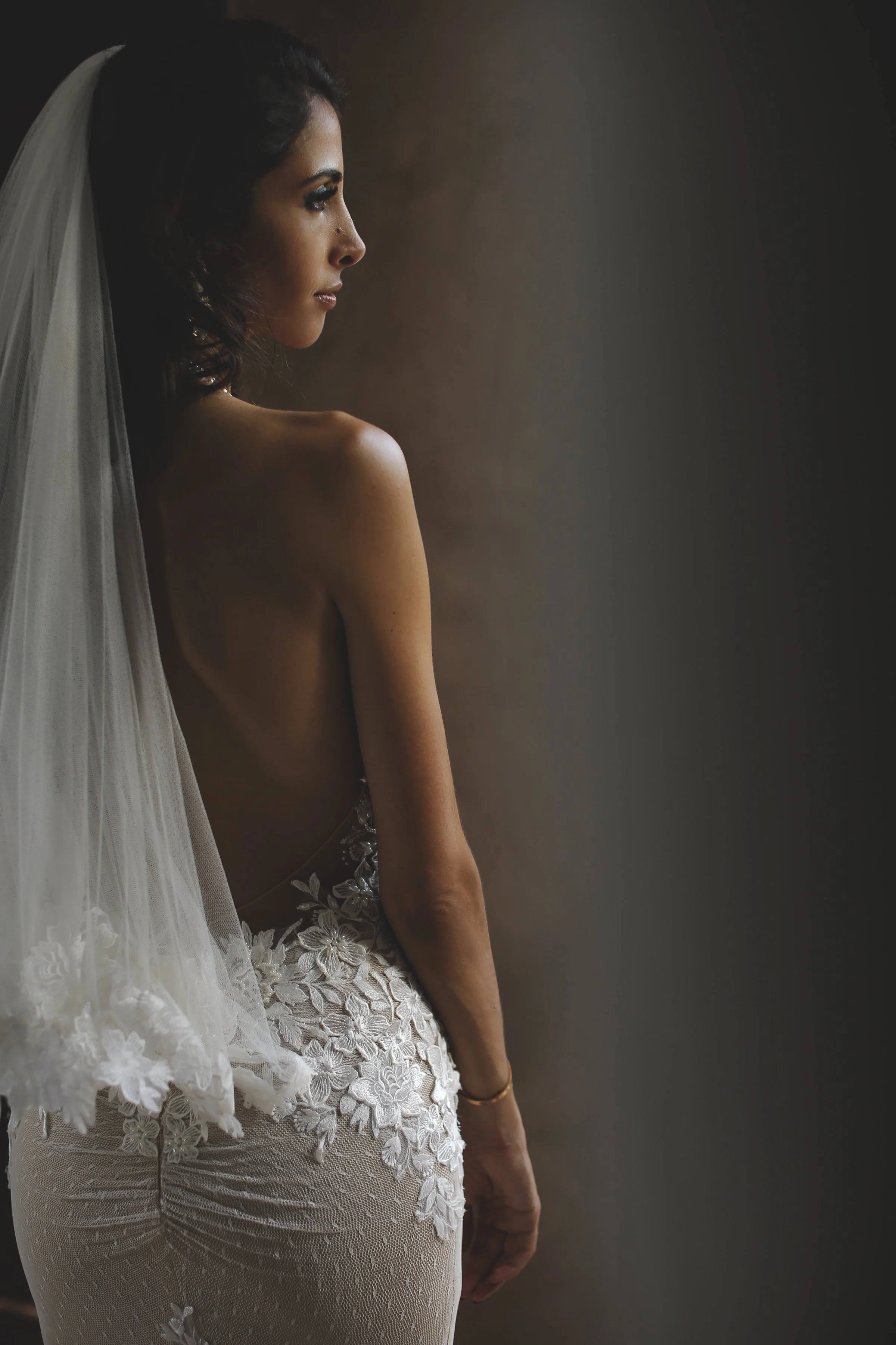 A woman with dark hair, wearing a wedding veil, looking to her left, partially dressed in a white lace wedding dress, standing against a dark background.