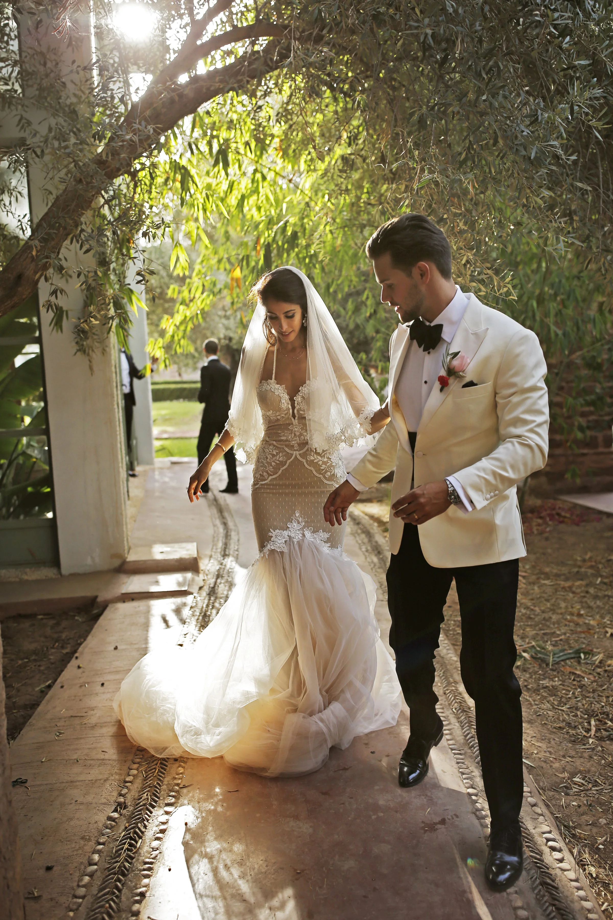 Bride in a lace wedding dress and veil walking with a groom in a white tuxedo under a tree canopy.