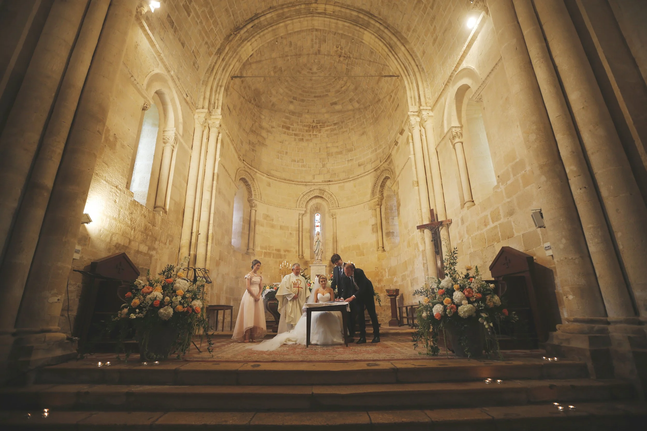 Wedding ceremony in a church with bride, groom, priest, and bridesmaids, surrounded by flower arrangements.