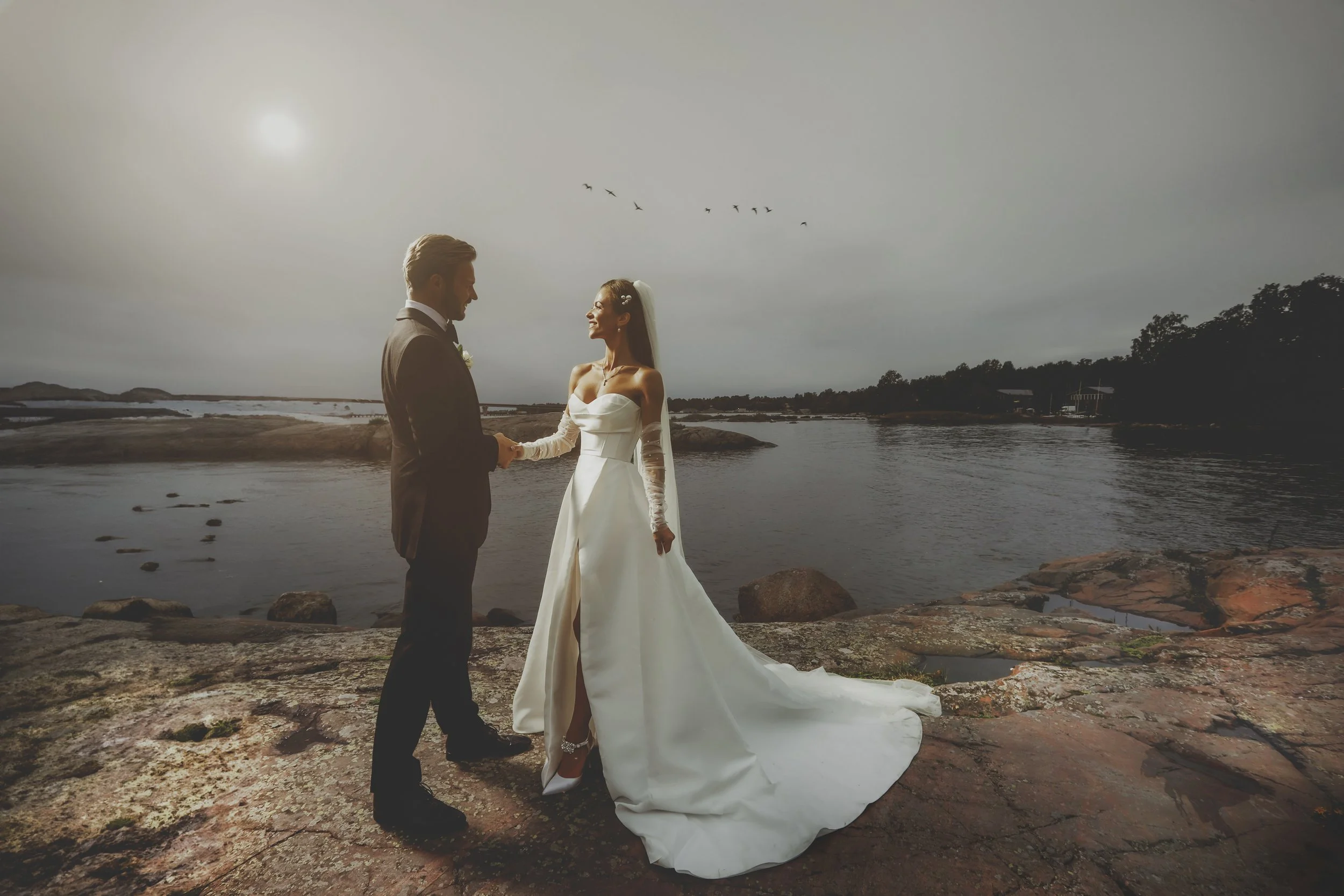 A bride and groom stand together on rocky terrain near water, holding hands and smiling at each other during a wedding ceremony, with birds flying in the cloudy sky above.