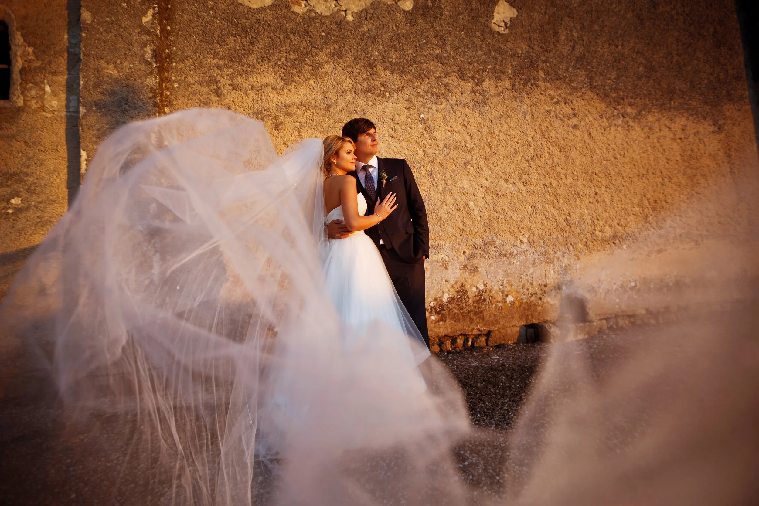 Bride and groom embracing near stone wall with flowing veil in foreground