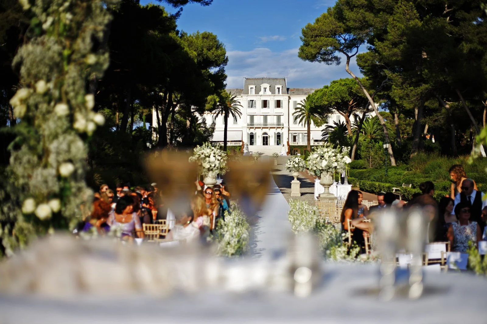 Outdoor wedding ceremony setup with chairs arranged on either side of an aisle decorated with white flowers, leading to a large white mansion surrounded by tall trees, with a clear blue sky overhead.