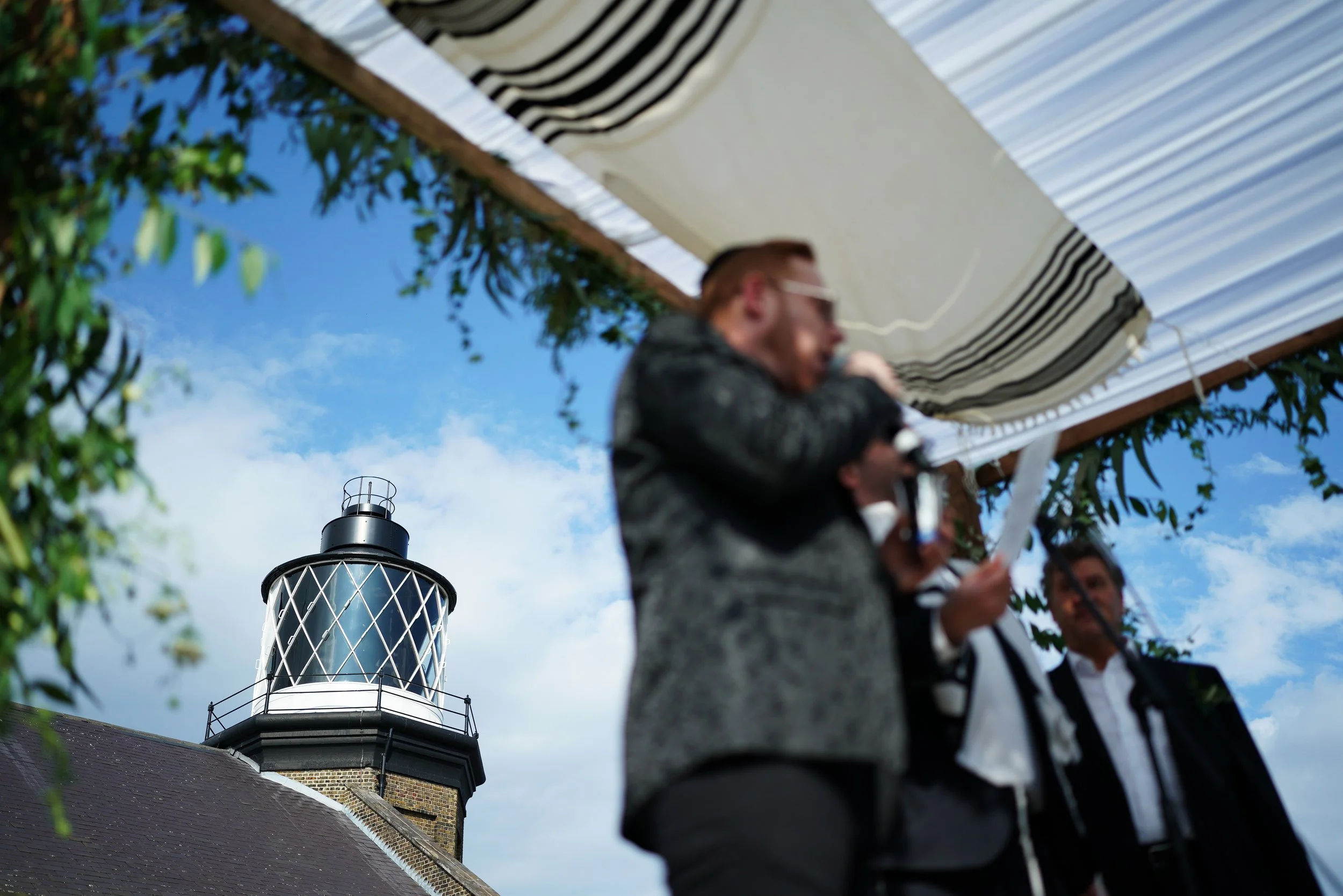 A blurry scene of three men at an outdoor event, with a lighthouse in the background under a partly cloudy sky.