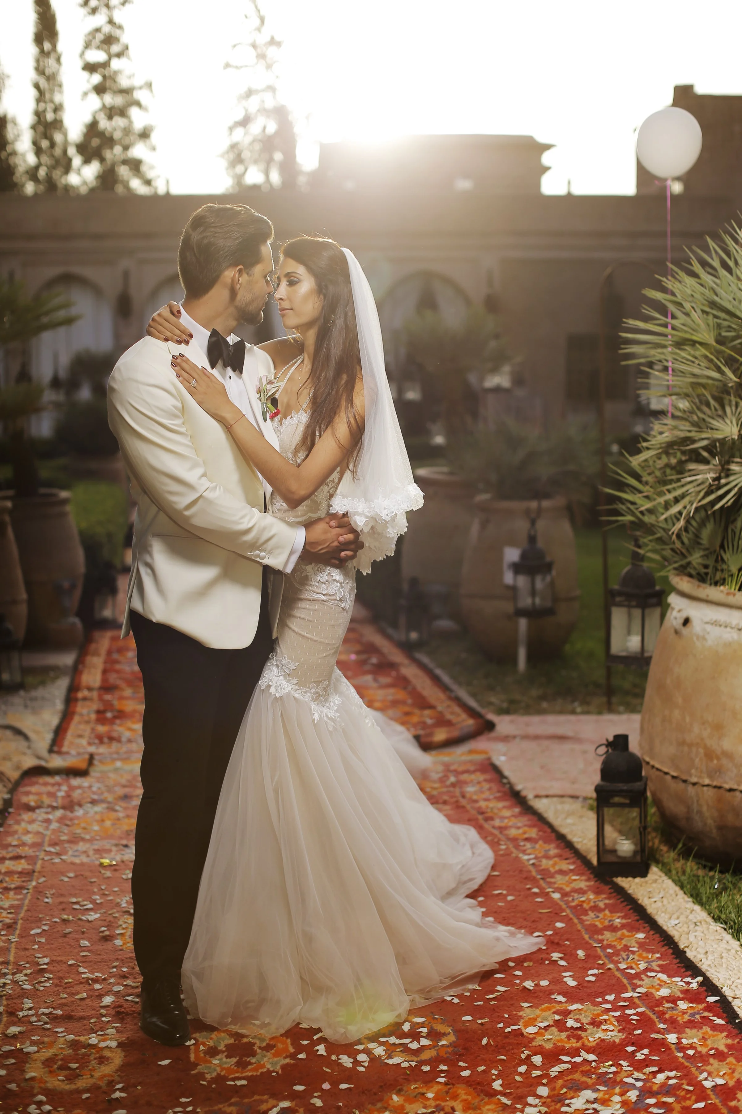 A bride and groom are dancing outdoors on a red carpeted area surrounded by petals at sunset. The groom is in a white tuxedo with a black bowtie, and the bride is in a white lace wedding gown with a veil. The background features a building with arche