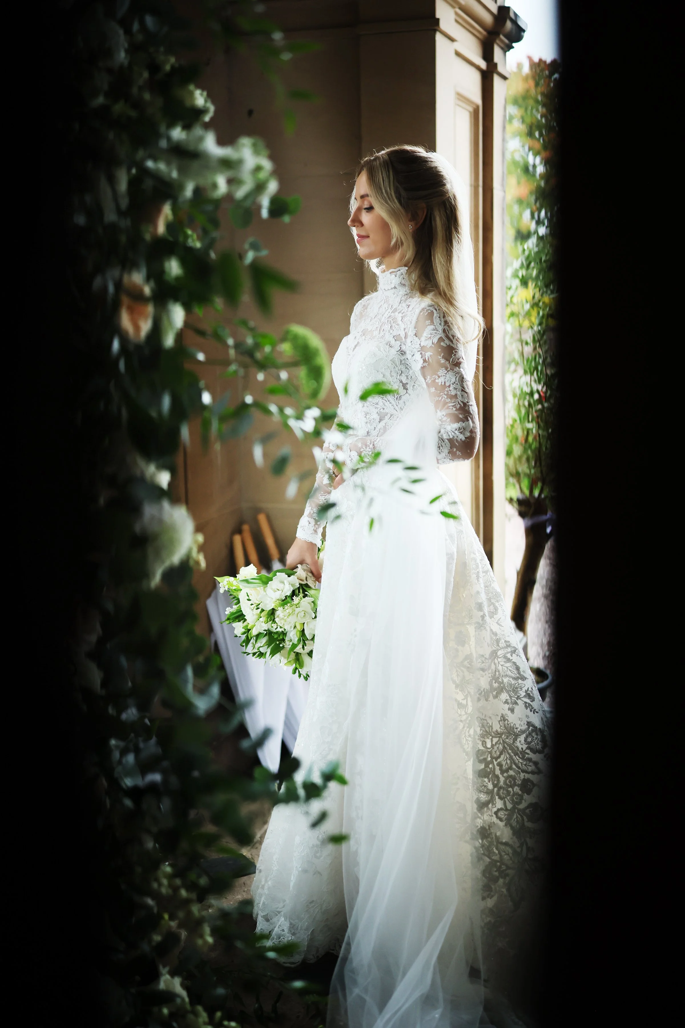 A bride in a white lace wedding dress, holding a bouquet of white and green flowers, standing near a window with sunlight streaming in, framed by greenery and plants.