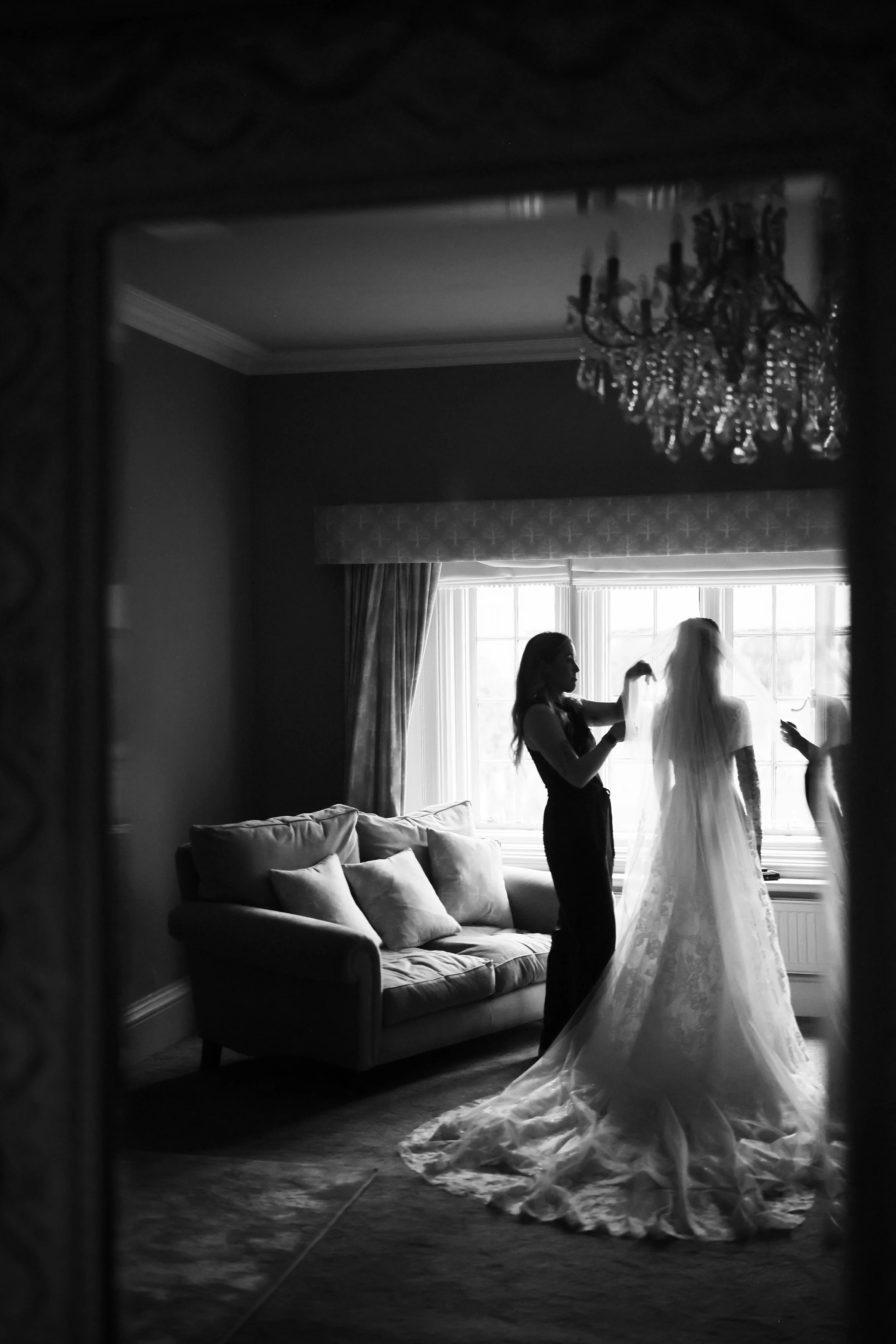 Looking through a mirror, a woman helps a bride with her wedding dress in a room with large windows, a chandelier, and a sofa.