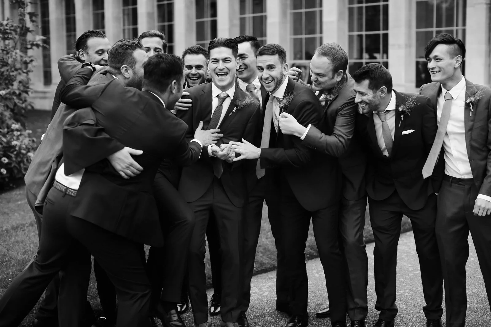 A group of men dressed in suits gathered outdoors, laughing and smiling together, celebrating at what appears to be a wedding or formal event.