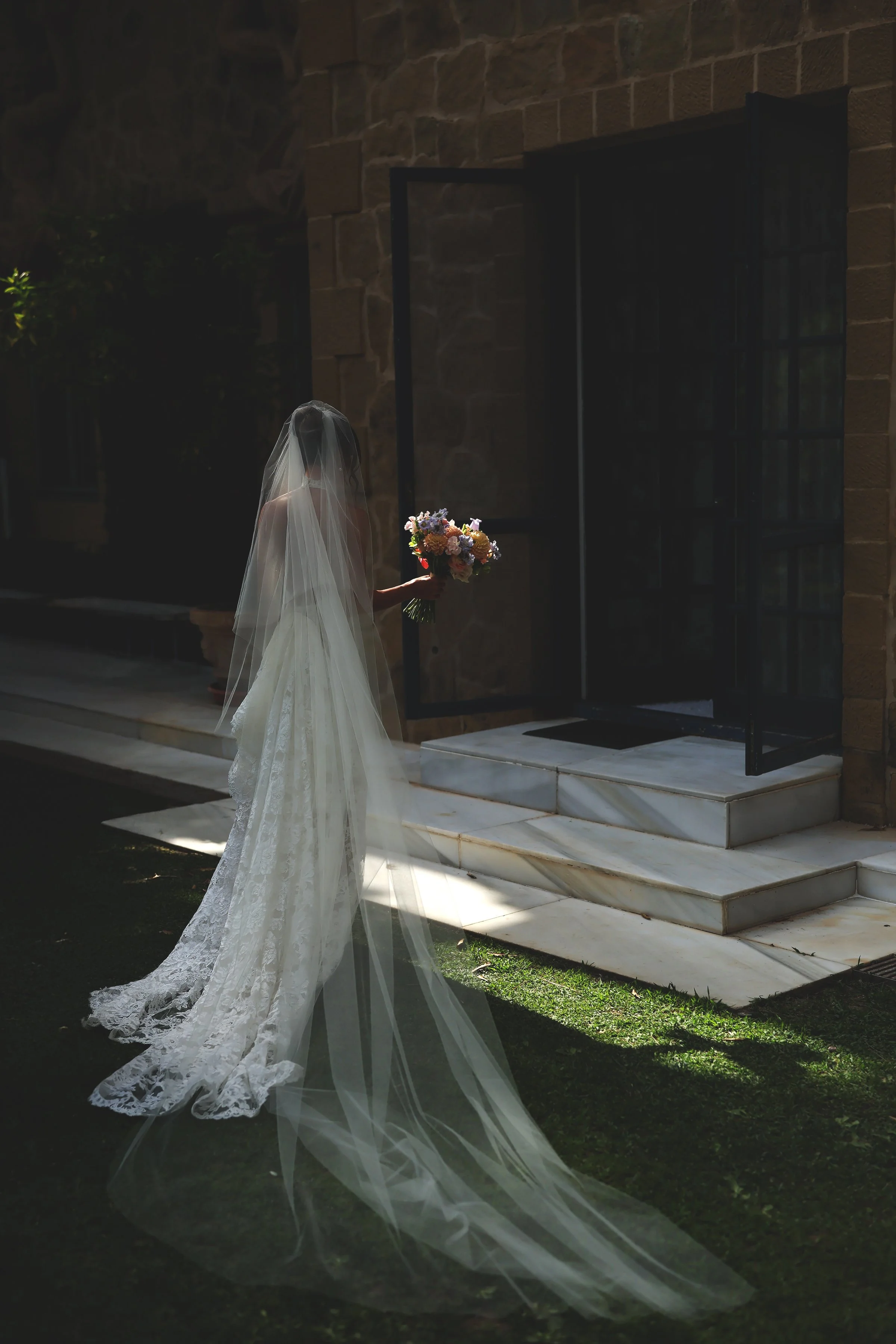 Bride wearing a white wedding dress with lace details and a long veil, holding a bouquet of flowers, standing outside near a building with stone walls and an open black door.