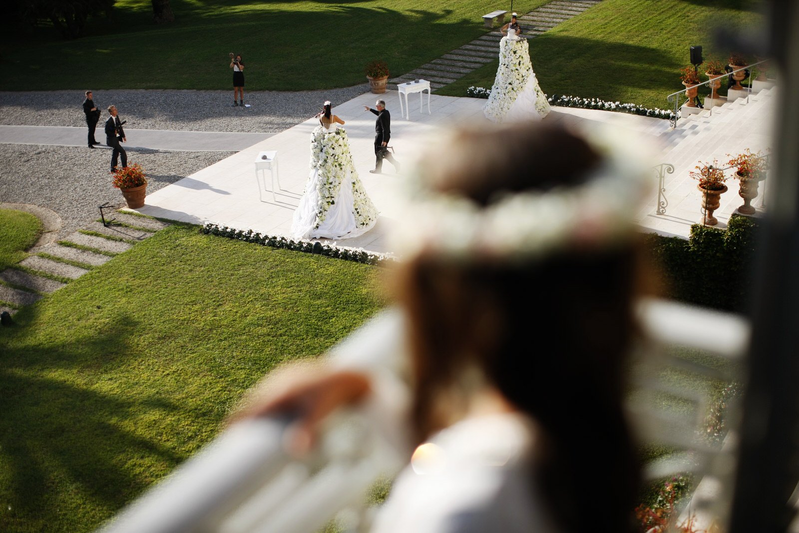 View of a wedding ceremony taking place outdoors, with a couple exchanging vows on a decorated platform, surrounded by guests and greenery, seen from a blurred foreground of a girl observing the event.
