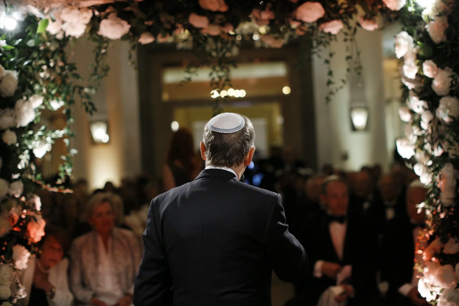 A man wearing a kippah and dark suit standing in front of an audience, during a formal ceremony or event at a decorated venue with floral arrangements.