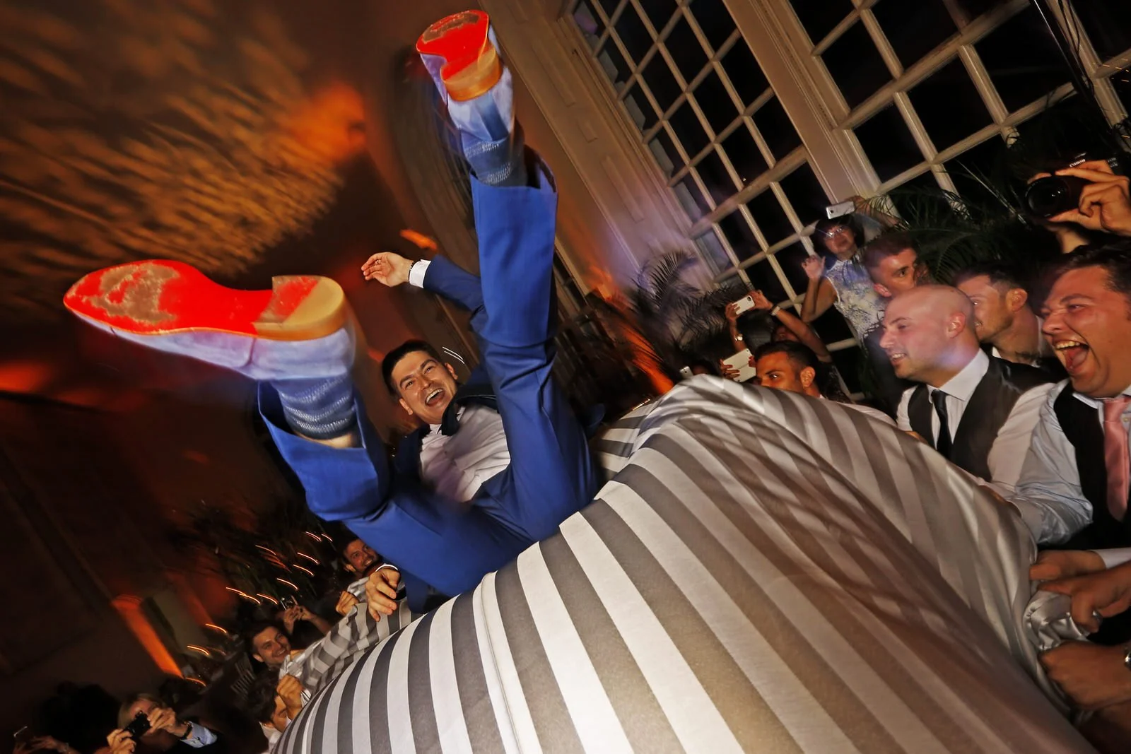 A celebration at a party where a man in a blue suit is being lifted upside down, showing his colorful shoes. Guests around are smiling and taking photos, with some people wearing formal attire.
